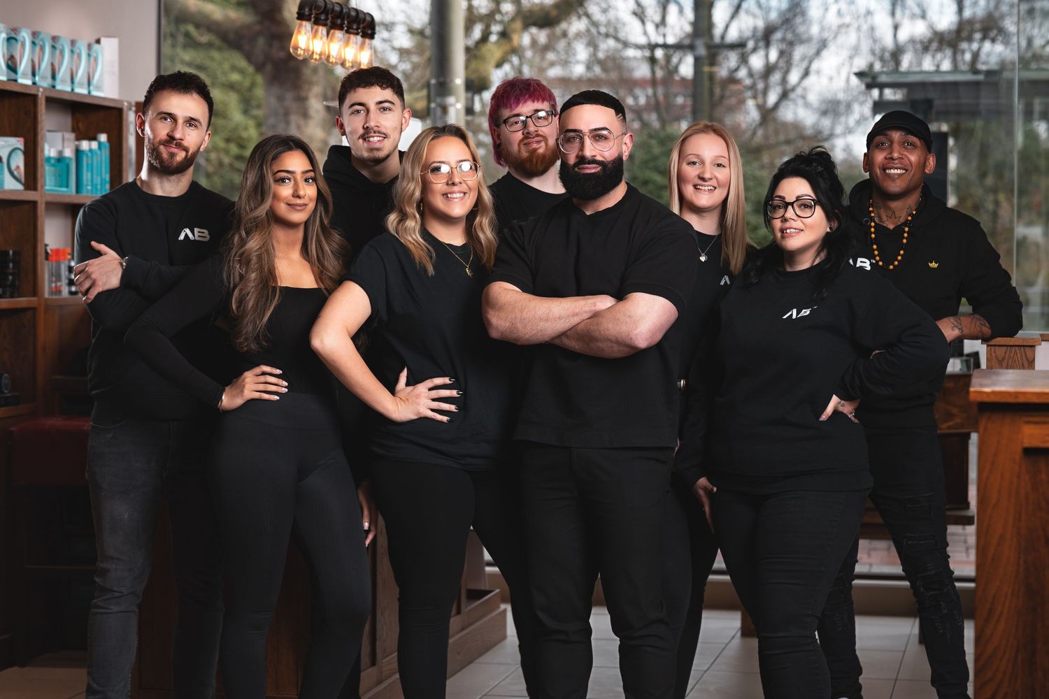 Nine-strong salon team in matching black branded outfits posed by the window under pendant bulbs at their Southampton barbershop, group team portrait photography