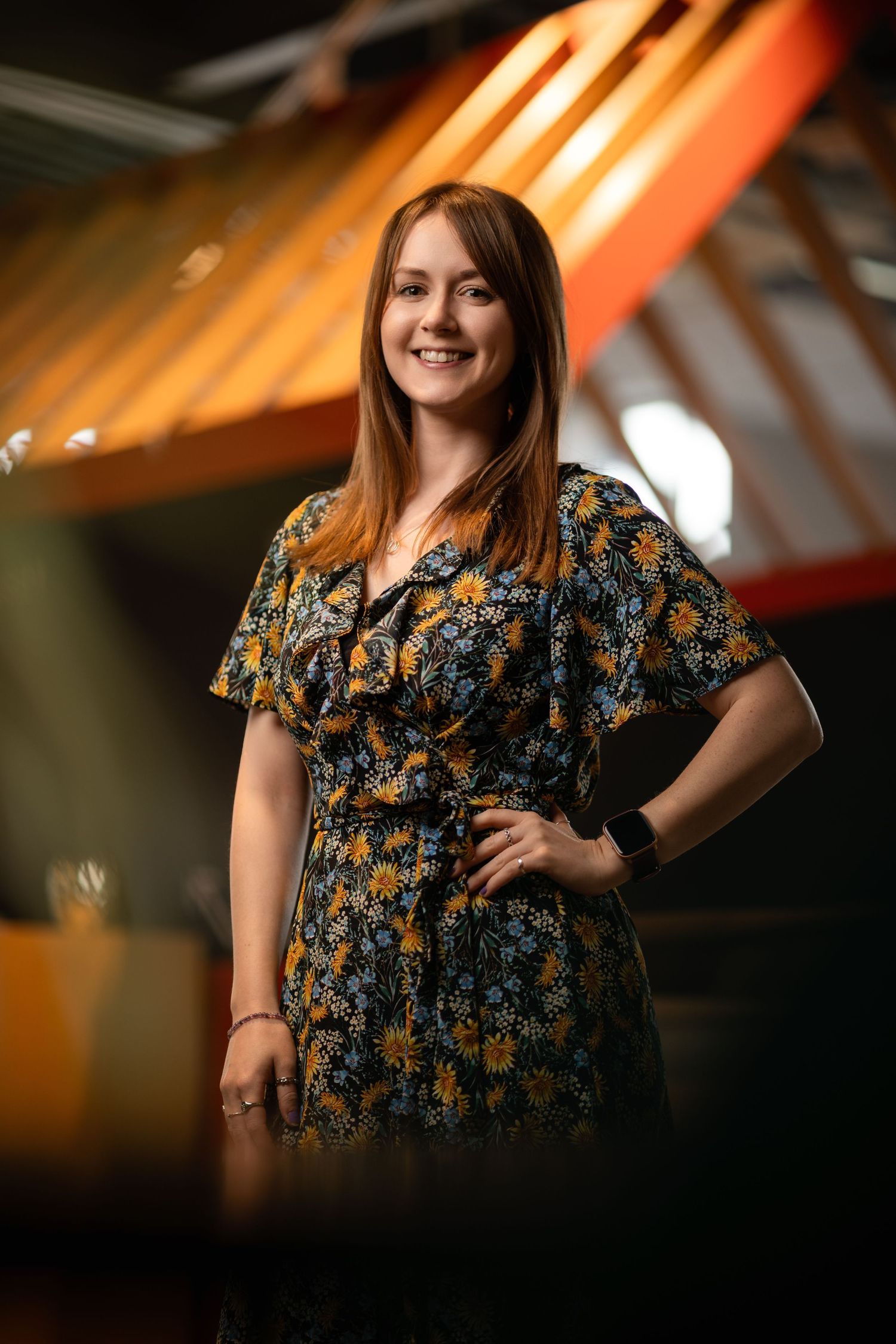 Smiling woman with auburn hair in a floral dress in a modern venue with warm overhead lighting, environmental business portrait photography
