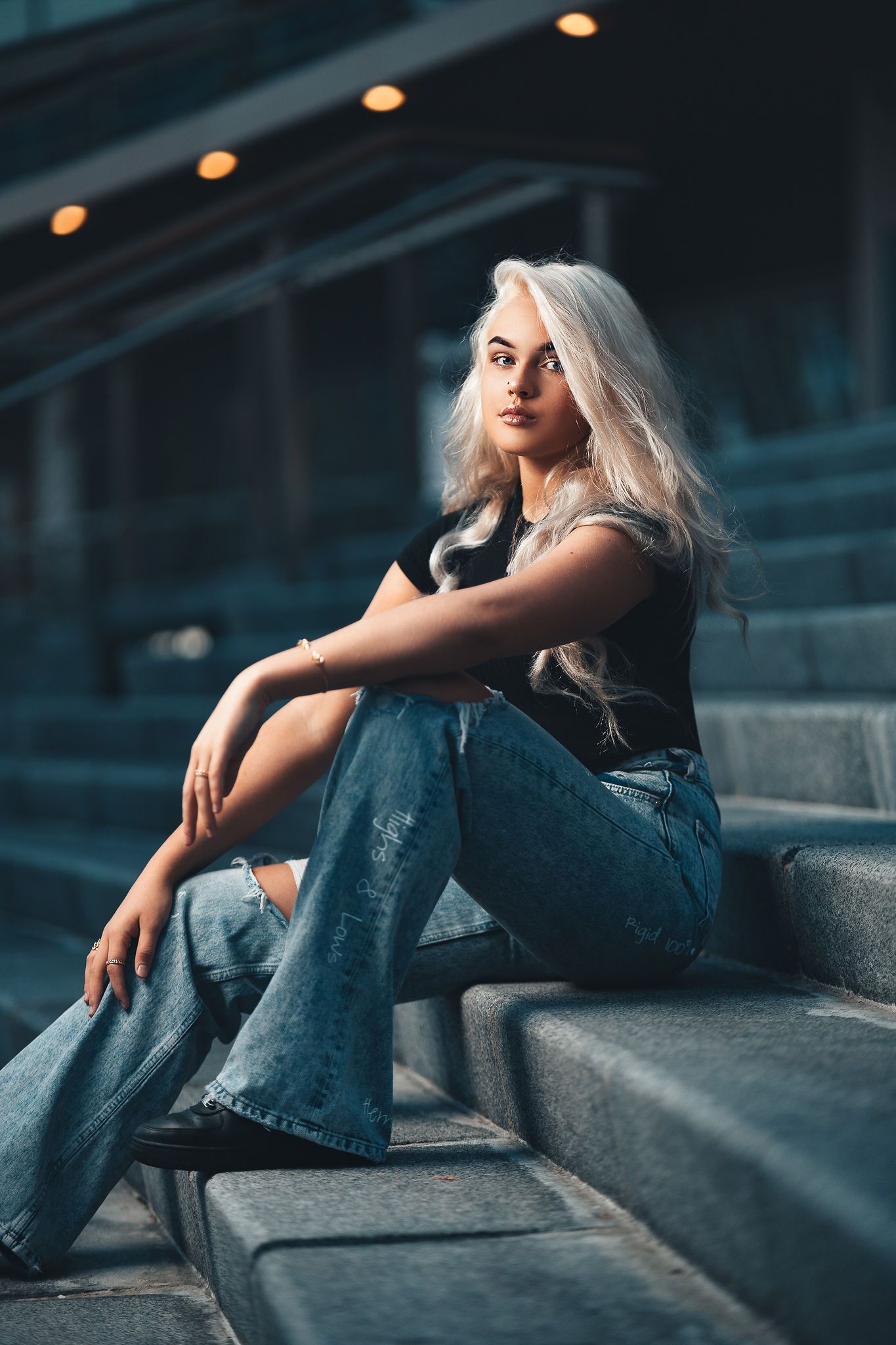 Amelia with platinum blonde hair in a black top and ripped jeans seated on concrete steps in blue-toned urban light, creative portrait photography Southampton