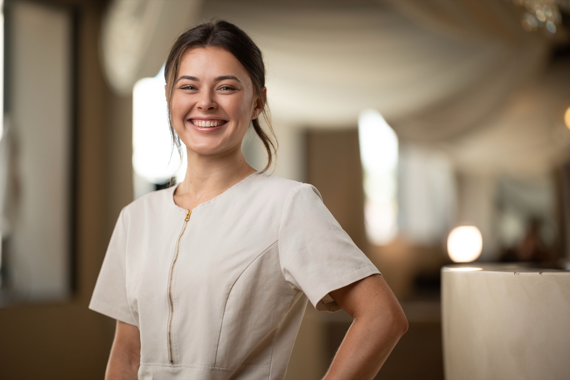 Beautician at Danica Payne smiling in a cream tunic with a hand on her hip in a bright salon interior, staff headshot photography
