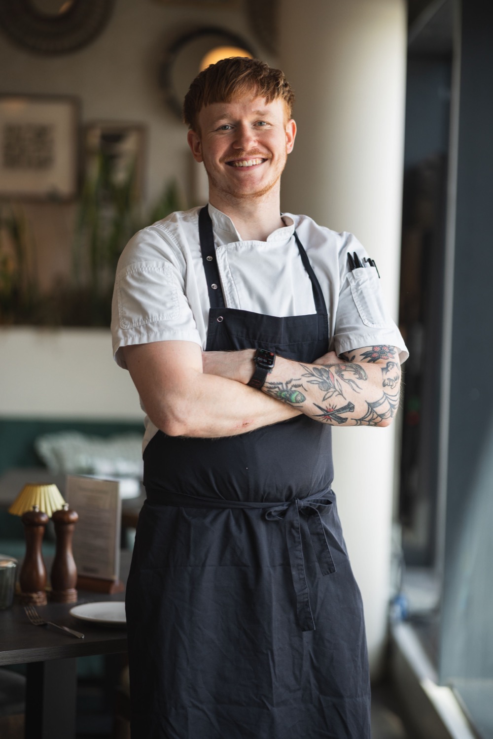 Sous chef in whites and dark apron with tattooed arms smiling in the restaurant at Figurati Italian restaurant Southampton, chef portrait photography