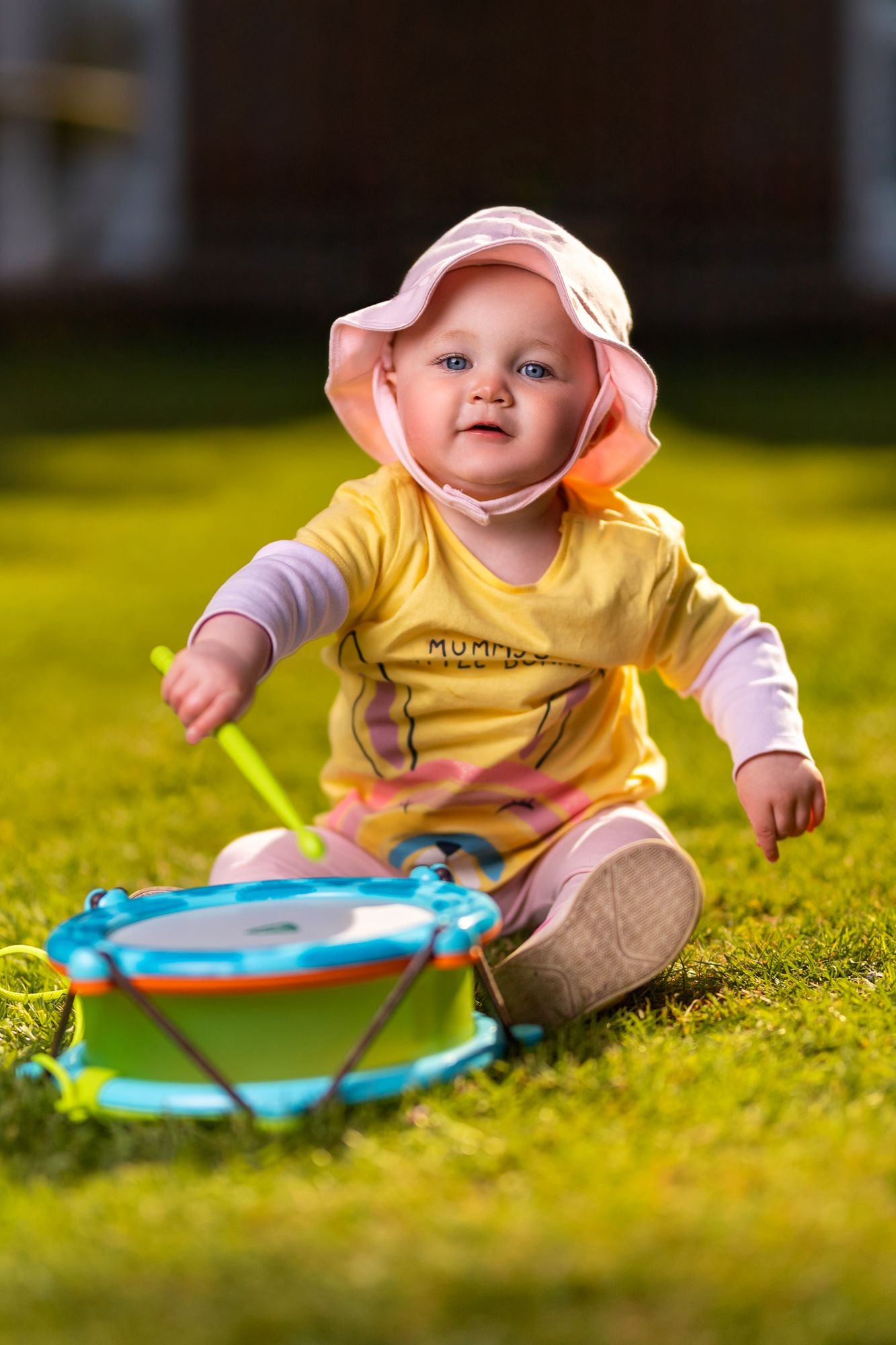 Baby in a yellow top and pink sun hat sitting on a sunny garden lawn playing with a toy drum, family portrait photography Southampton