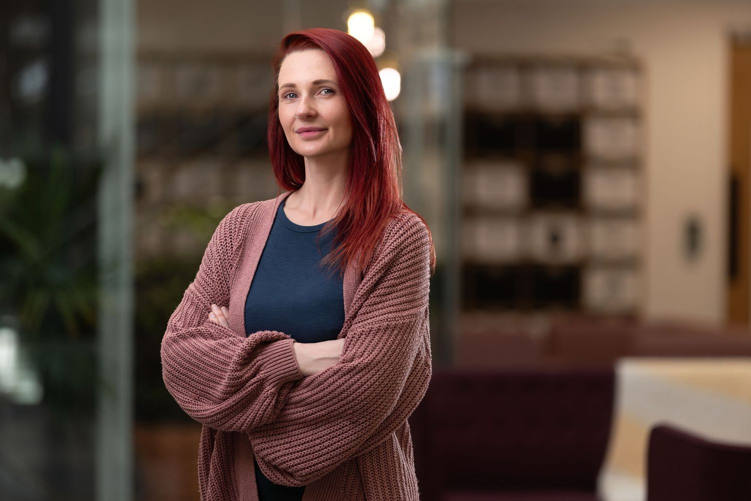 Grace with red hair in a pink knitted cardigan and navy top, arms crossed in a modern office interior with glass partitions, relaxed professional portrait photography Southampton