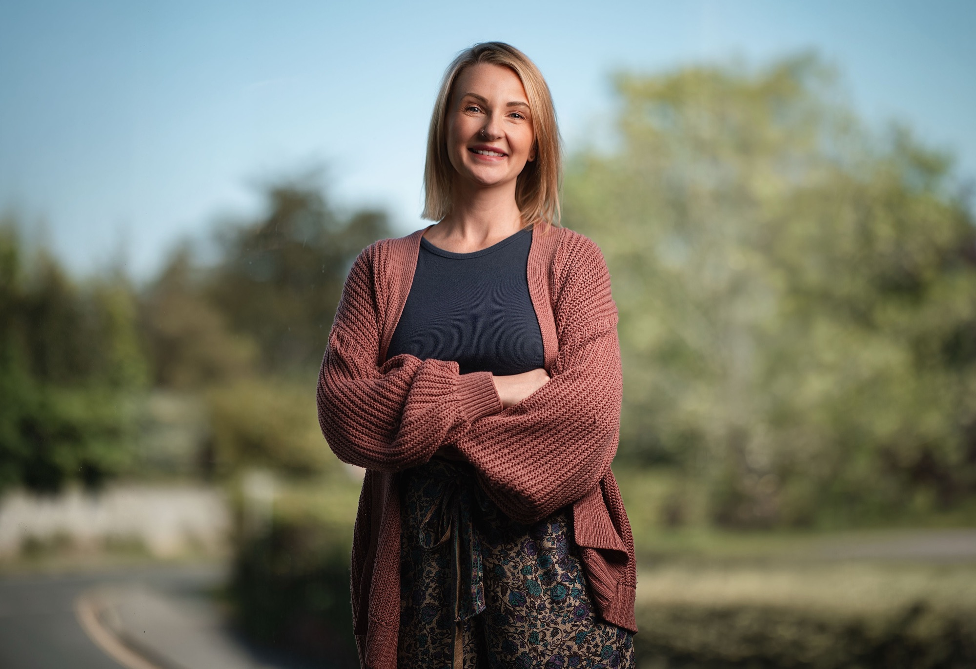 Therapist smiling in a rust cardigan with arms crossed outdoors in soft natural light, personal branding headshot photography Hampshire