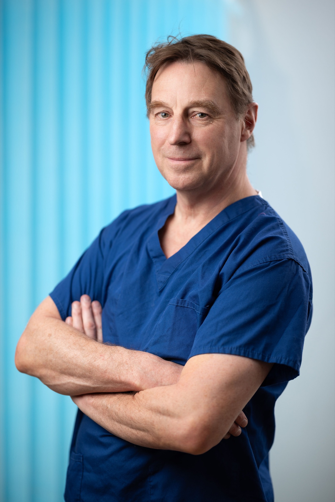 Mr Nigel Horlock, breast surgeon at Hampshire Breast Clinic, in blue surgical scrubs with arms crossed, professional medical headshot photography
