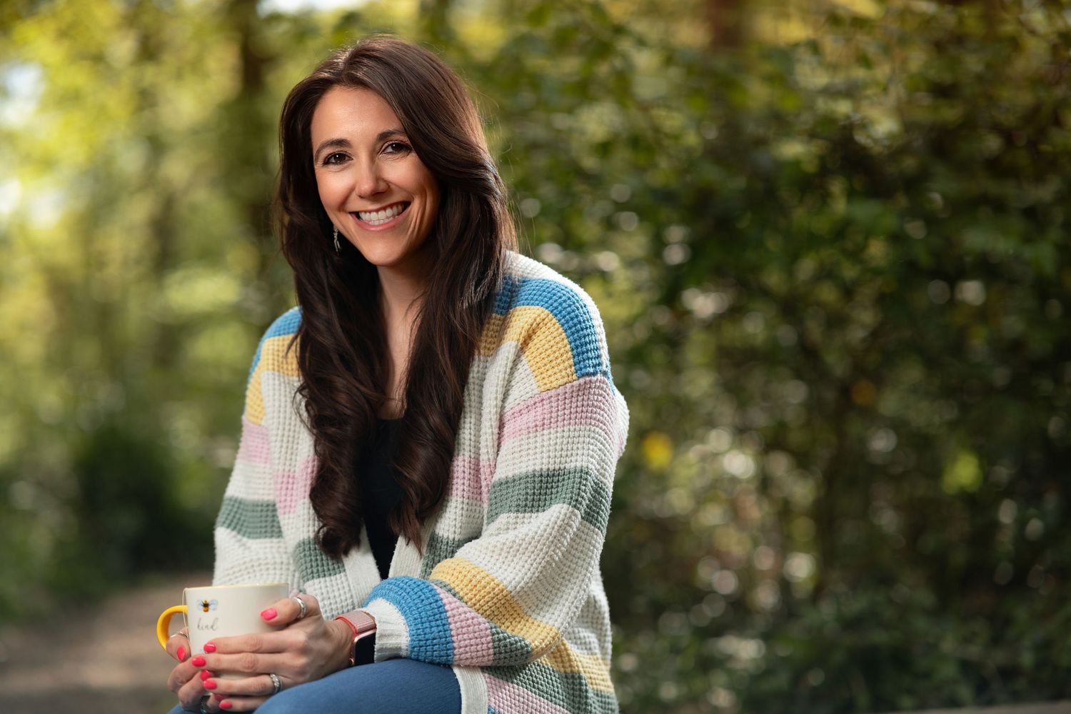 Holly smiling with long dark hair in a colourful striped cardigan holding a mug, seated outdoors with green bokeh background, portrait photography