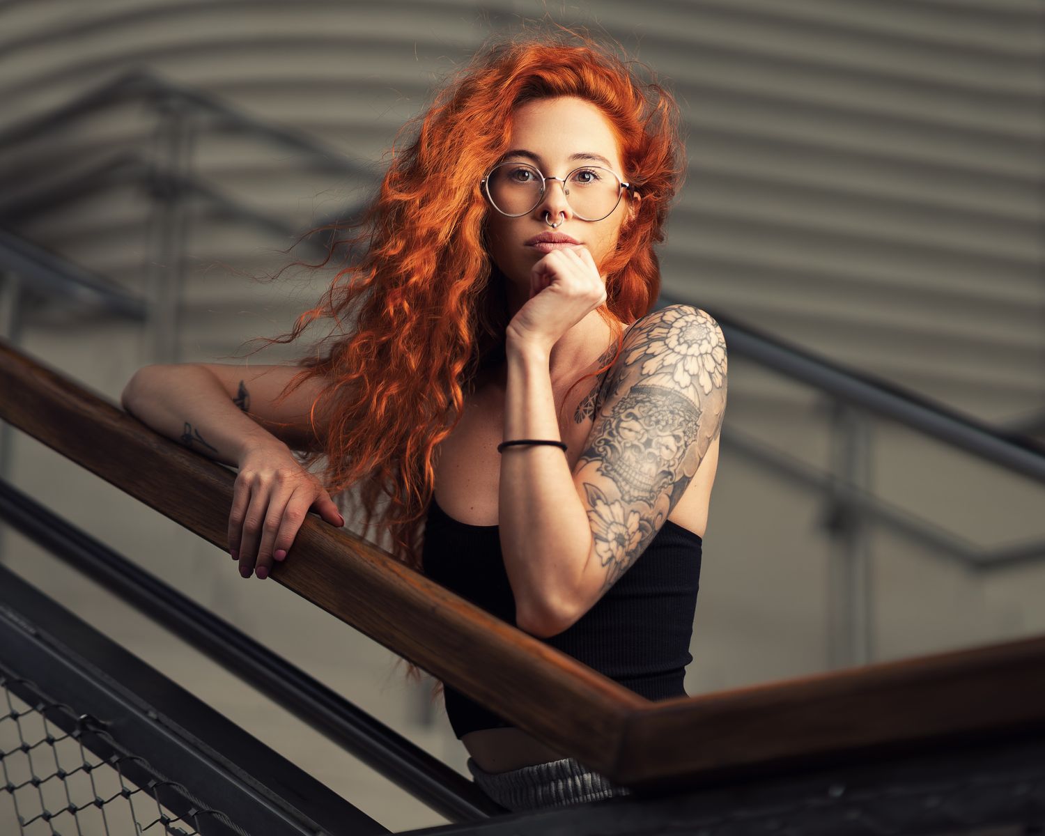 Kadie with red curly hair, glasses and tattoo sleeve leaning on a metal staircase railing in a black vest top, alternative portrait photography Southampton