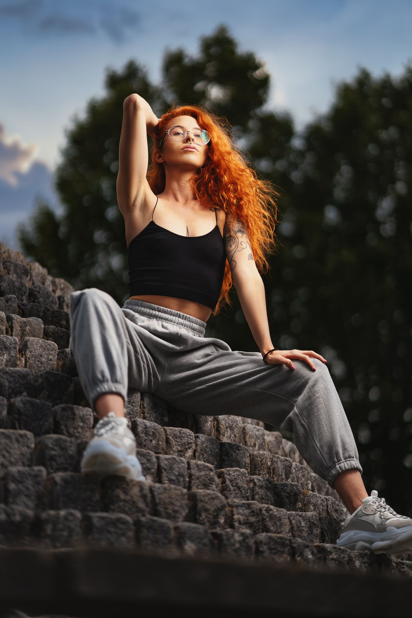 Kadie with long red curly hair and glasses seated on stone steps in a black crop top and grey joggers with tattoo sleeve, outdoor portrait photography