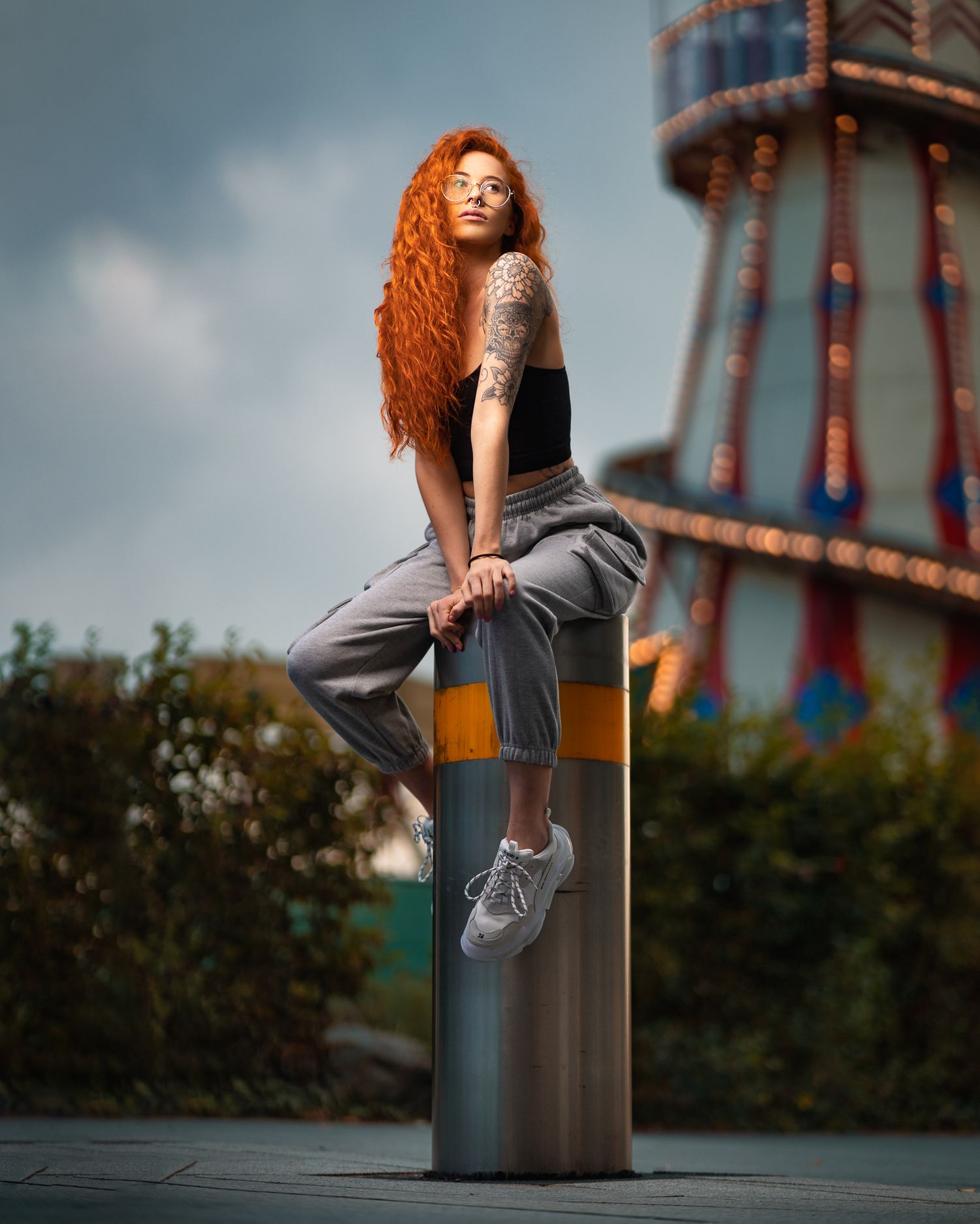 Kadie with long red curly hair, glasses and tattoo sleeve perched on a metal bollard with a fairground helter skelter behind, creative outdoor portrait photography Southampton