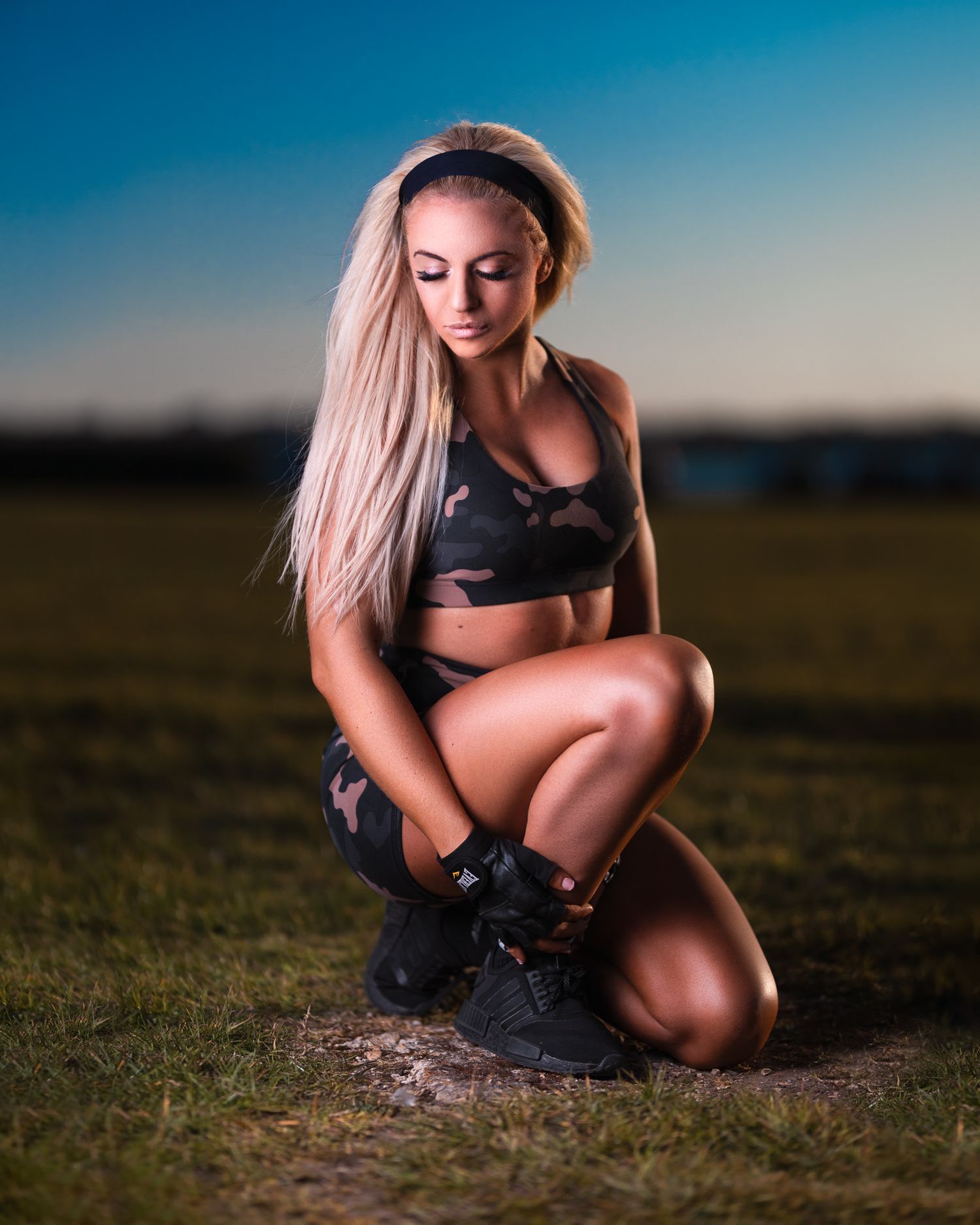 Kyra with long blonde hair in camo print sportswear crouching on grass at dusk with dramatic sky, fitness portrait photography Southampton