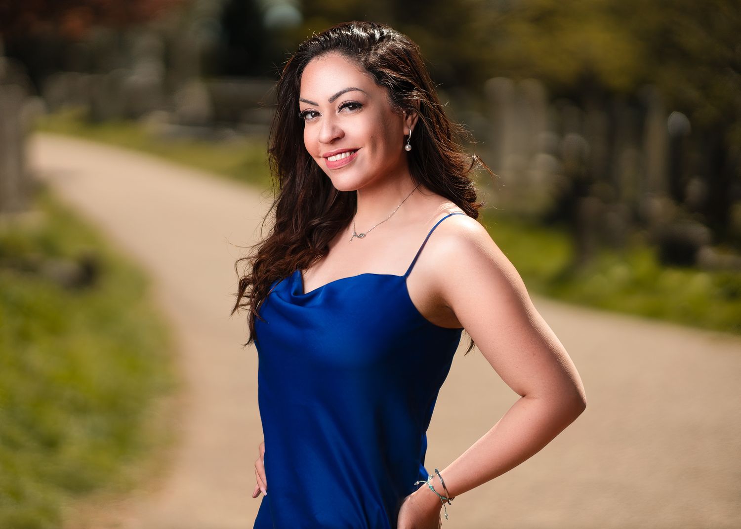 Leilah with long dark curly hair in a blue satin slip dress smiling on a park path in soft outdoor light, portrait photography Southampton