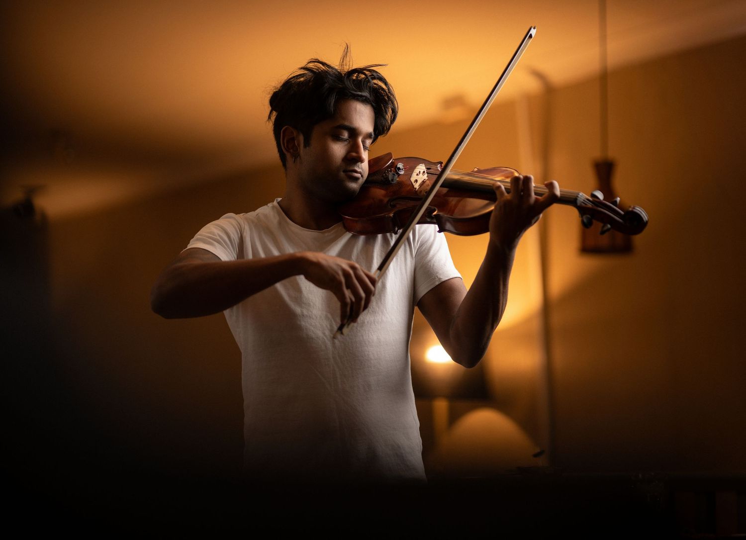 Mahesh in a white t-shirt playing violin with eyes closed in warm amber lighting, creative musician portrait photography Southampton