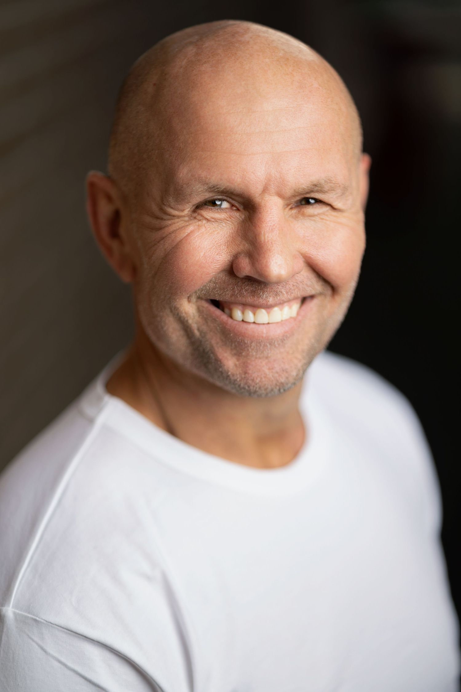 Smiling bald man in a white t-shirt, natural-light headshot with a warm bokeh background, personal branding photography