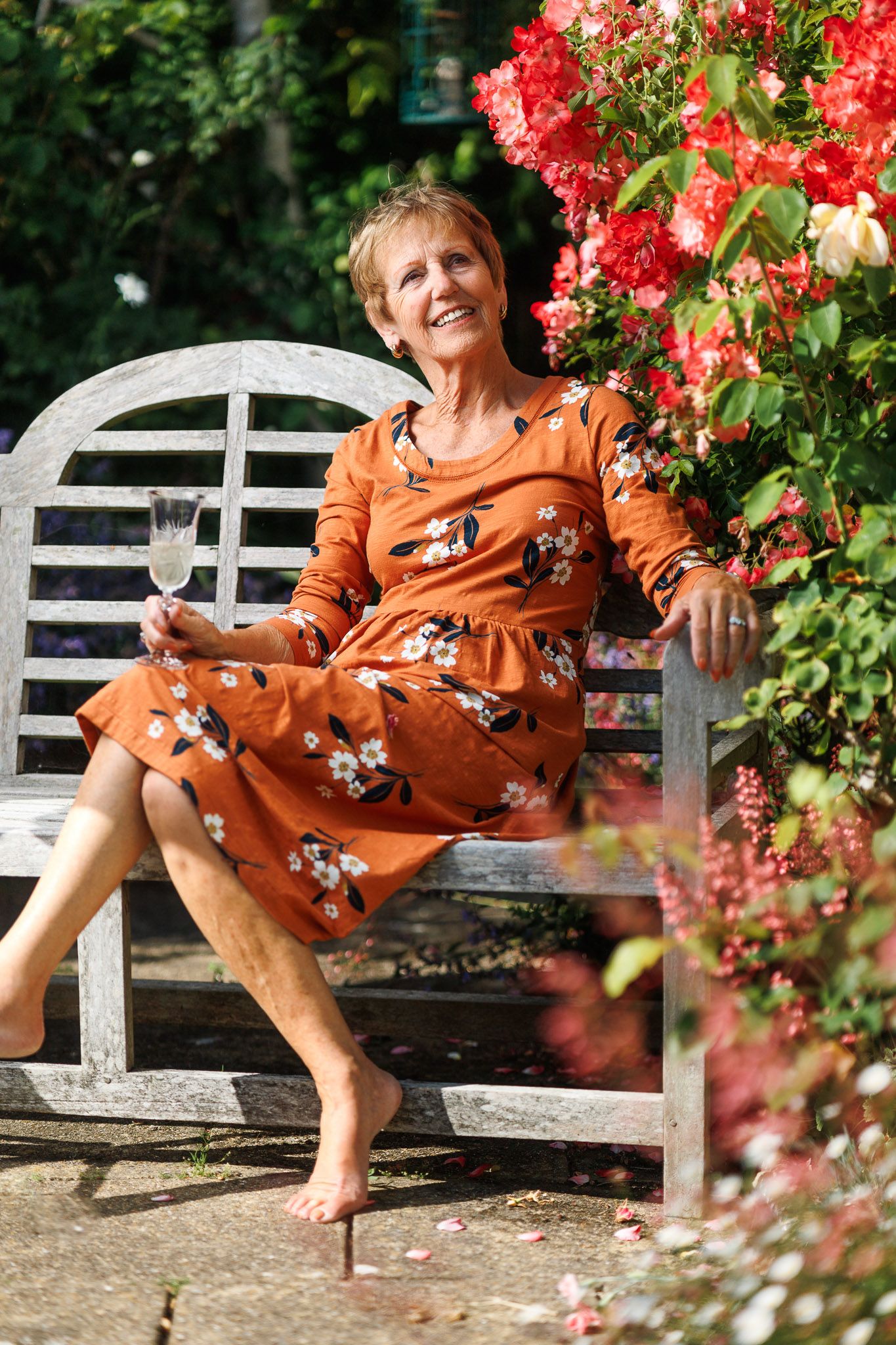 Rosemary smiling in an orange floral dress seated on a wooden garden bench beside red climbing roses with a glass of wine, outdoor portrait photography