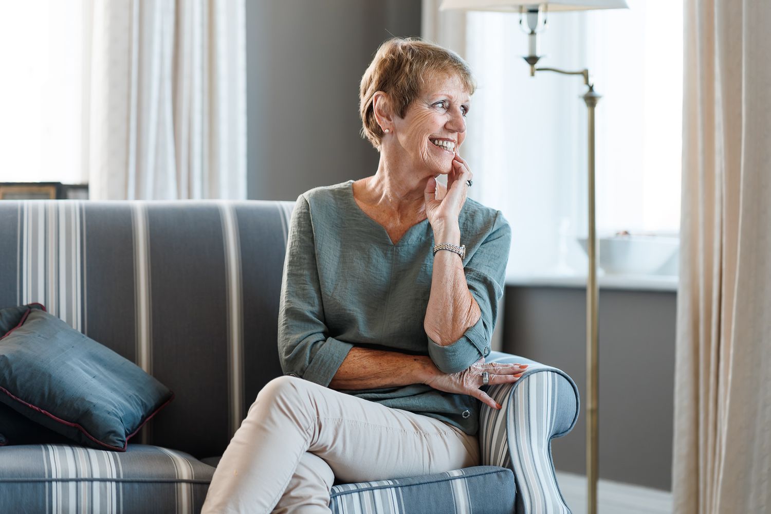 Rosemary smiling in a sage green top seated on a striped armchair in a bright living room by a window, relaxed indoor portrait photography Southampton