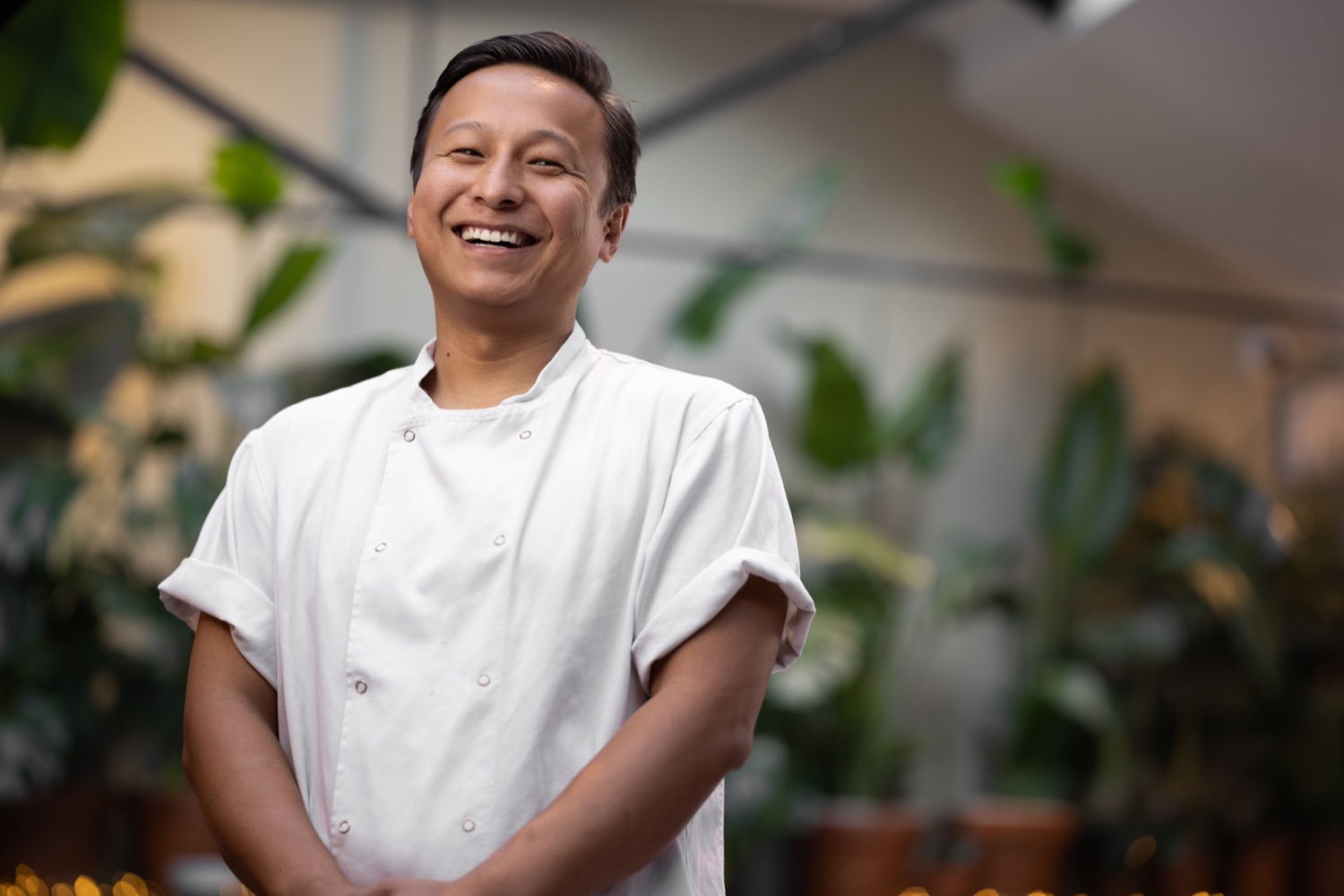 Smiling chef in white kitchen jacket surrounded by tropical houseplants at The Mayfair Chippy Knightsbridge, chef portrait photography London