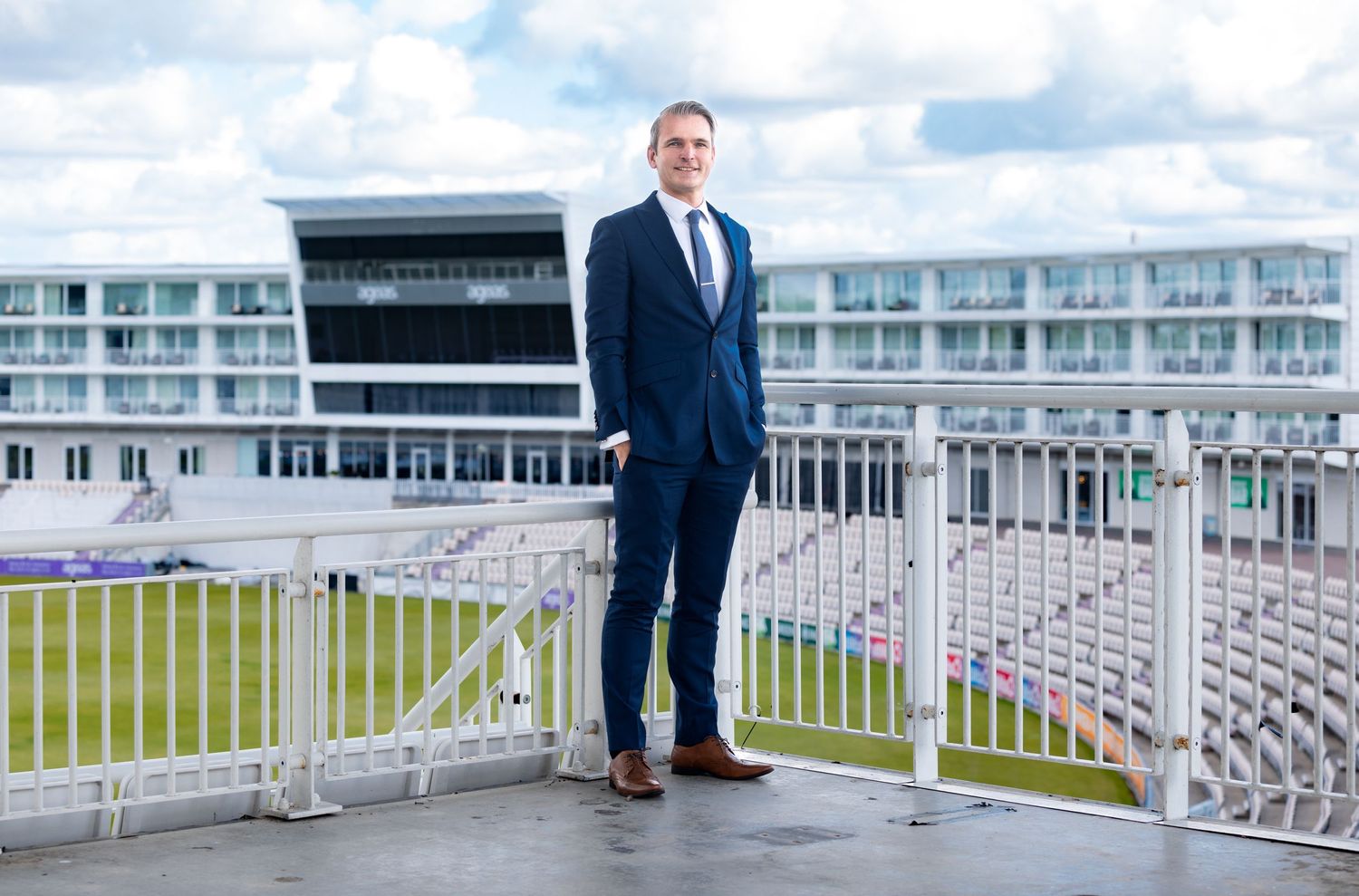 Steven Exley in a navy suit standing on a balcony overlooking a cricket ground with stadium building behind, environmental business portrait photography Southampton