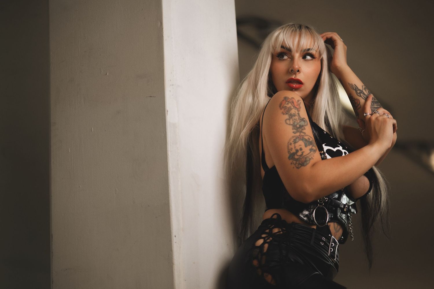 Tattooed woman with platinum blonde hair in a black PVC outfit leaning against a pillar in an industrial space, alternative fashion portrait photography Southampton