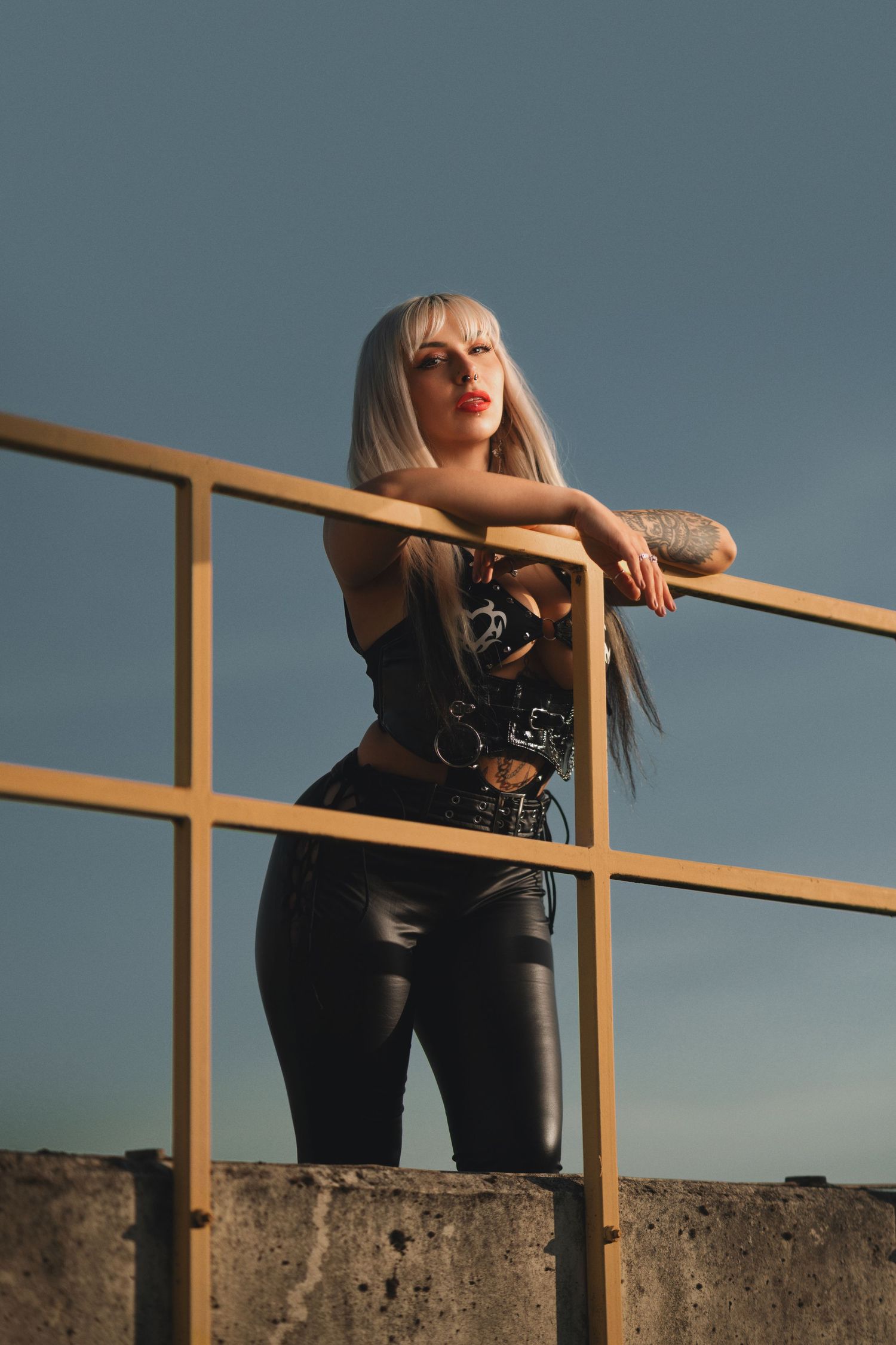Tattooed woman with platinum blonde hair leaning on a yellow metal railing in a black harness top and leather trousers, alternative fashion portrait photography