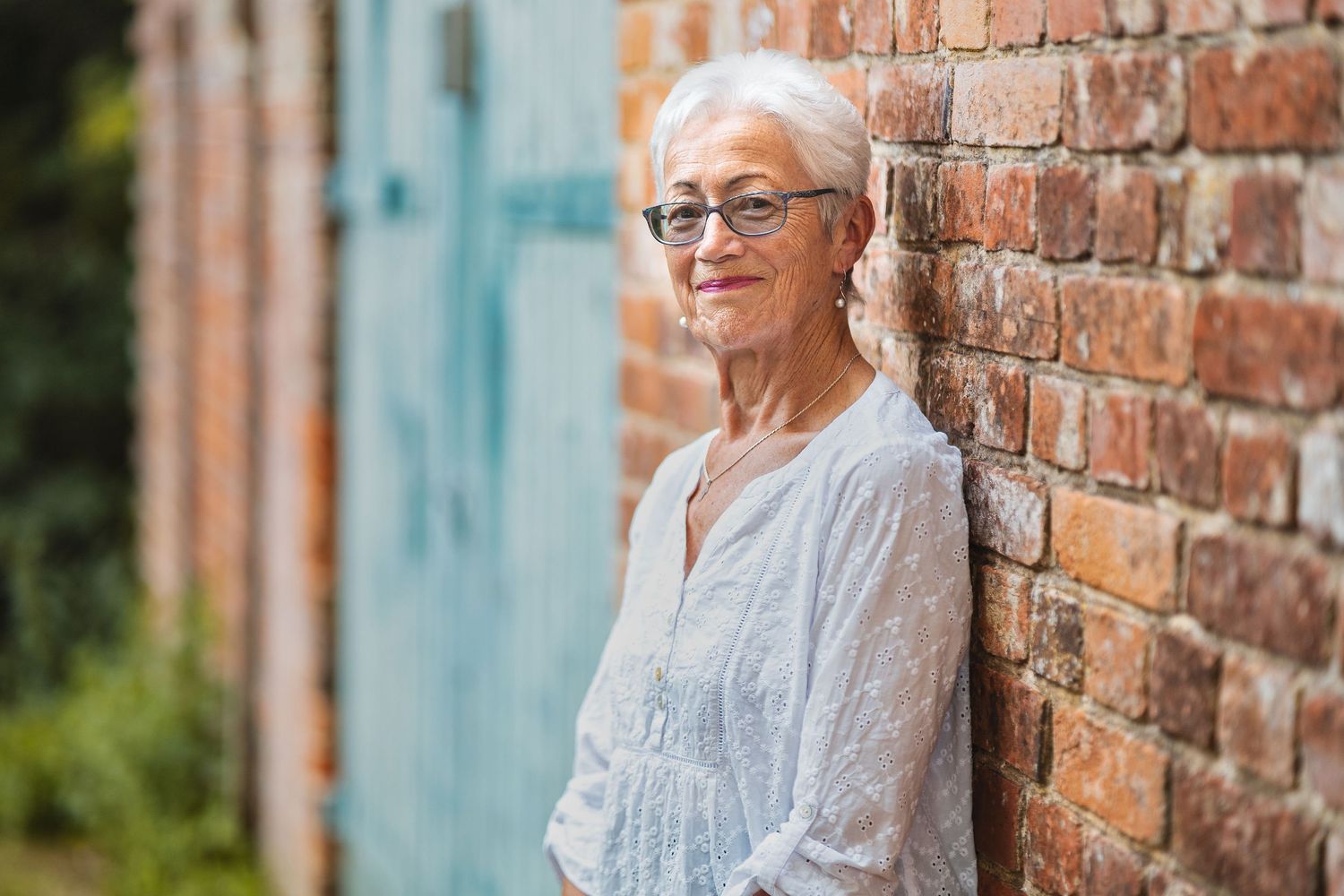 Sue with short white hair and glasses in a white broderie anglaise blouse leaning against a red brick wall with a blue door, outdoor portrait photography Southampton