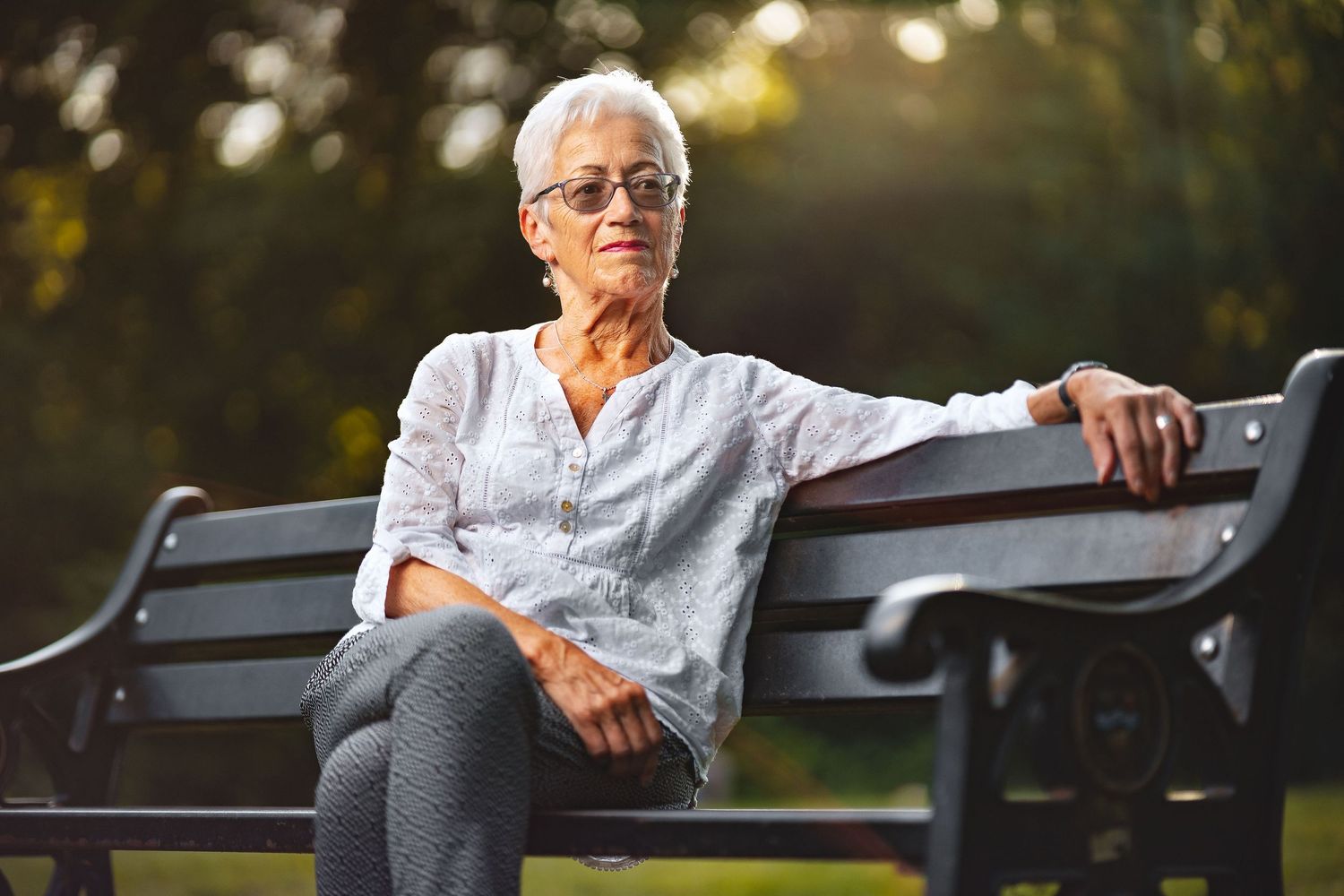 Sue with short white hair and glasses in a white broderie anglaise blouse seated on a park bench in warm evening light, relaxed outdoor portrait photography Southampton