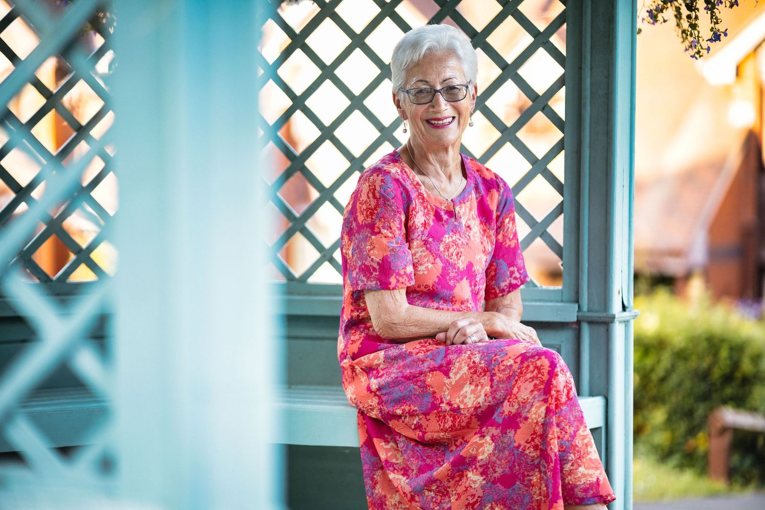 Sue with short grey hair and glasses in a pink floral dress smiling in a painted garden gazebo, outdoor lifestyle portrait photography Southampton