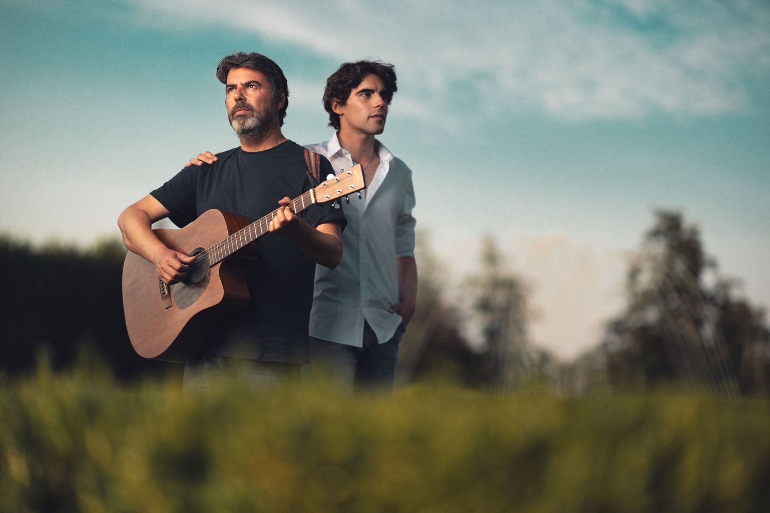 Father and son duo outdoors with acoustic guitar, standing in long grass against a dramatic sky at golden hour, musician portrait photography Southampton
