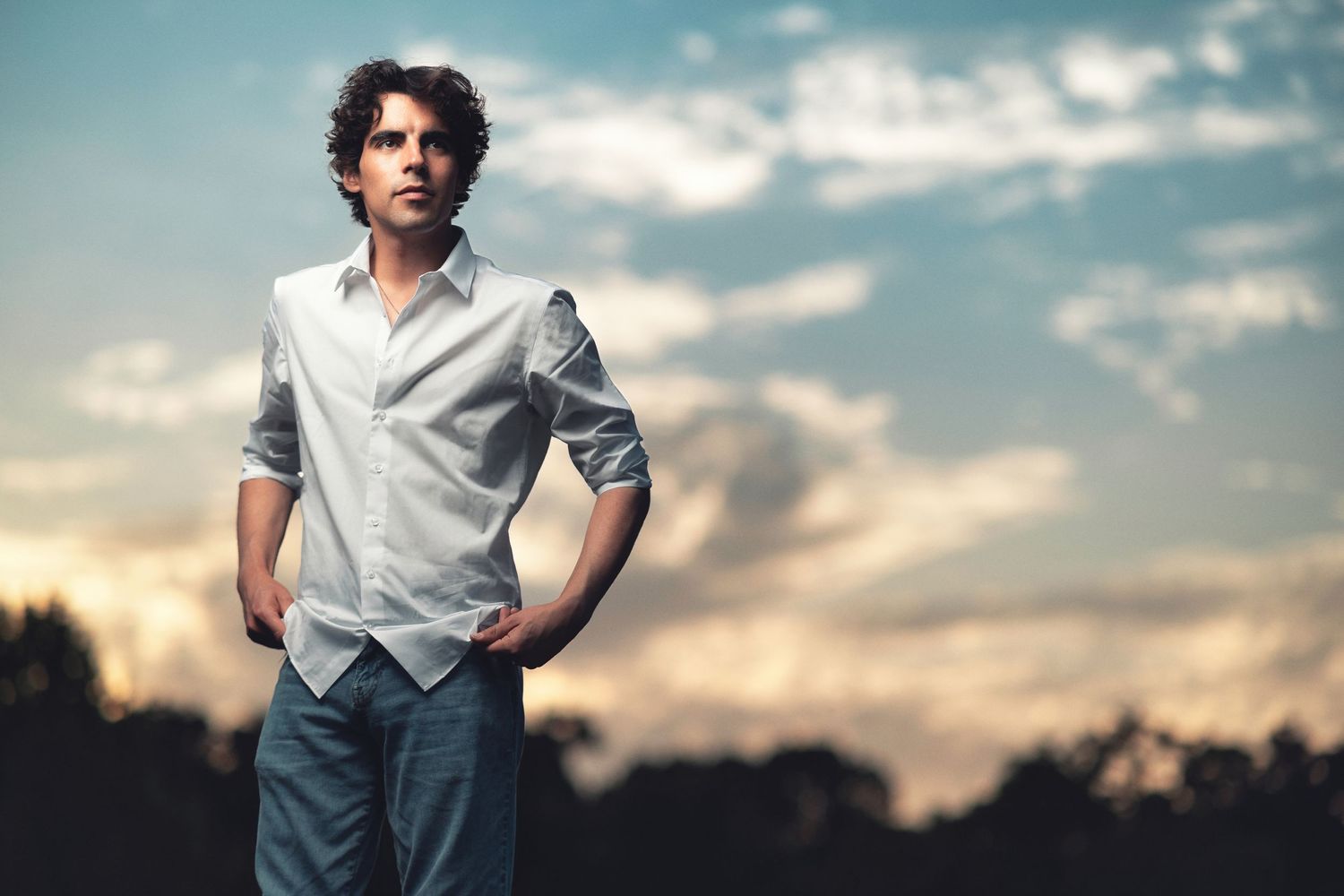 Young man with curly dark hair in an open white shirt and jeans, hands in pockets against a dramatic sunset sky, portrait photography