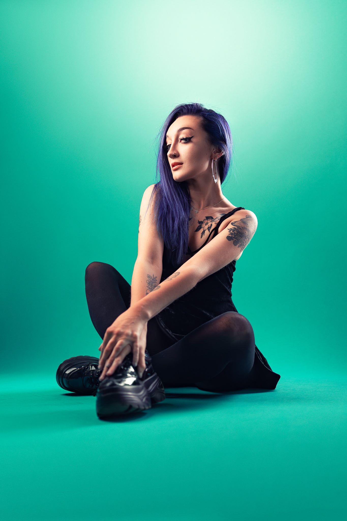 Tattooed woman with purple hair in a black outfit seated cross-legged on a turquoise studio backdrop, creative studio portrait photography Southampton