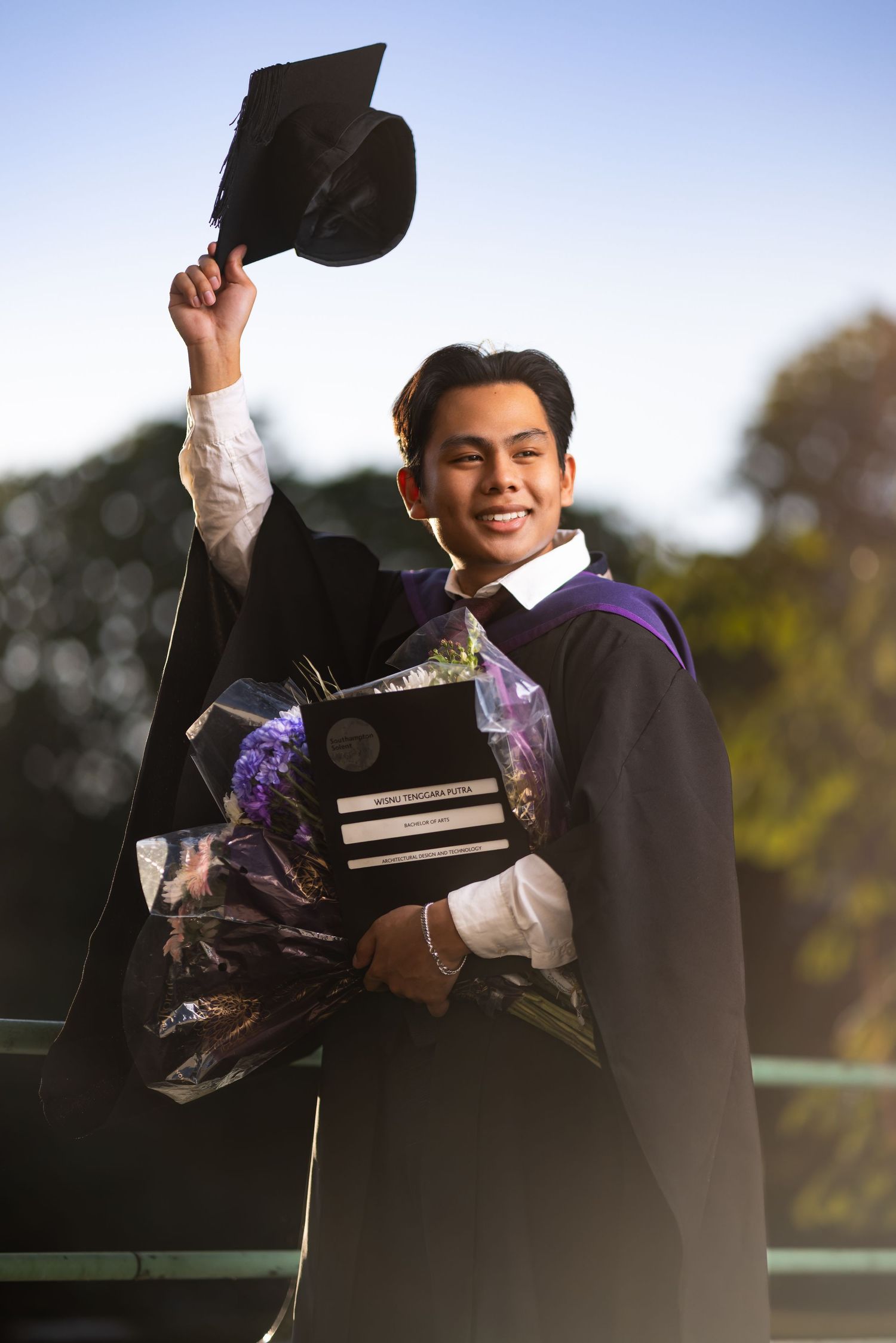 Wisnu in graduation robes holding up mortarboard cap with bouquet and degree certificate, outdoor graduation portrait photography