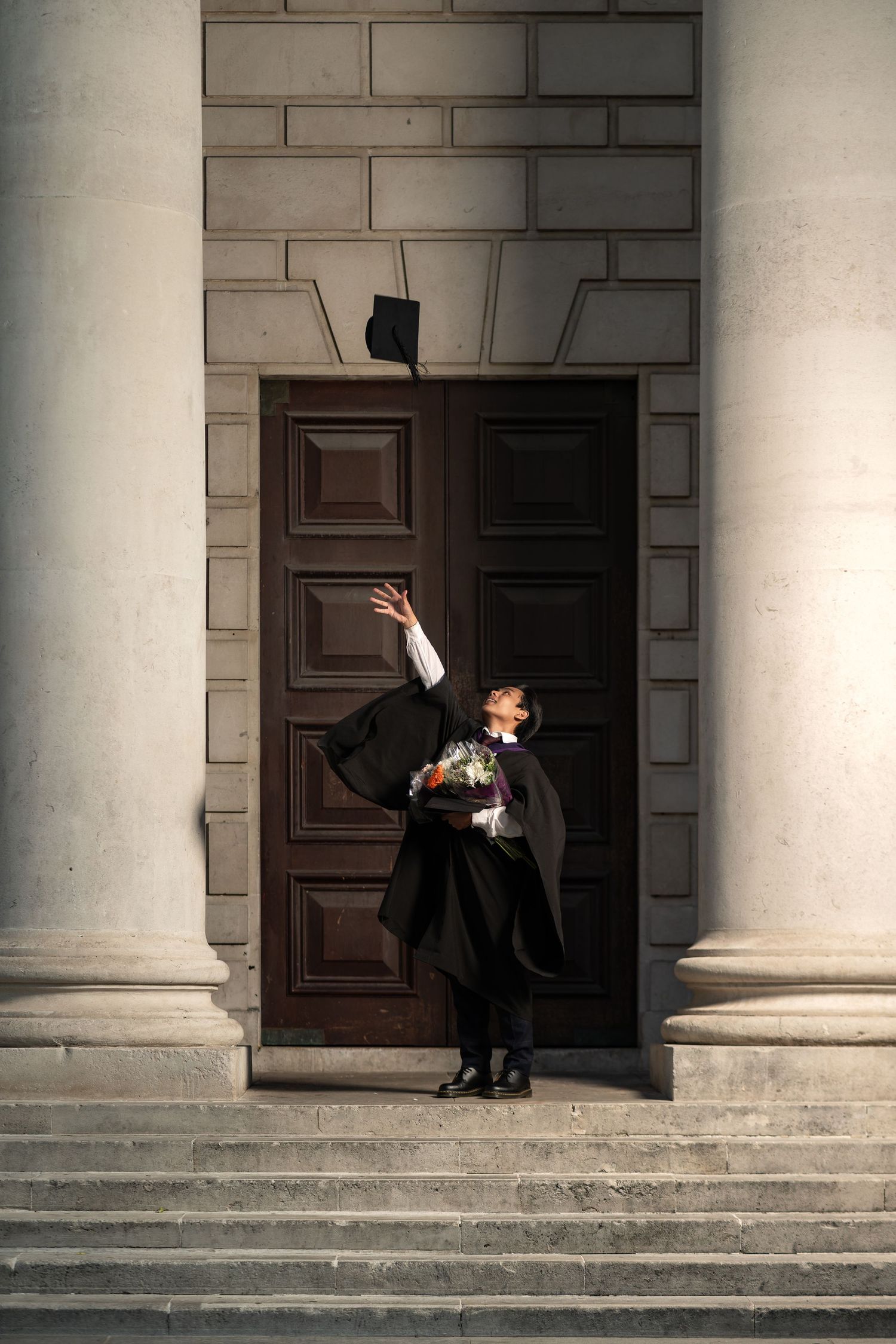 Wisnu in graduation robes tossing mortarboard cap in the air outside a grand columned stone building, graduation portrait photography Southampton
