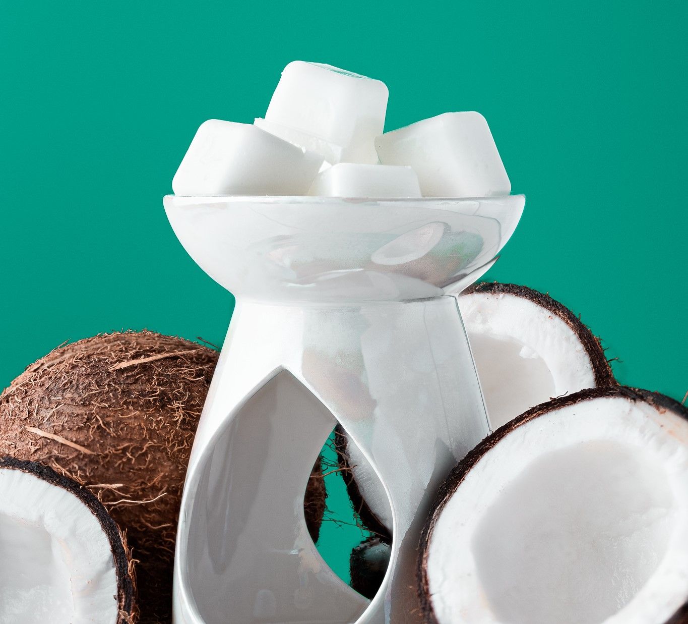 White coconut wax melts in a ceramic burner surrounded by whole and halved coconuts on a teal background, home fragrance product photography Southampton