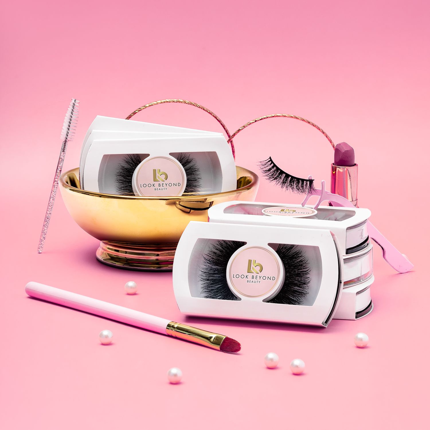 Look Beyond Beauty false eyelash sets styled with makeup brushes, lipstick and pearls in a gold bowl on a pink background, beauty product photography