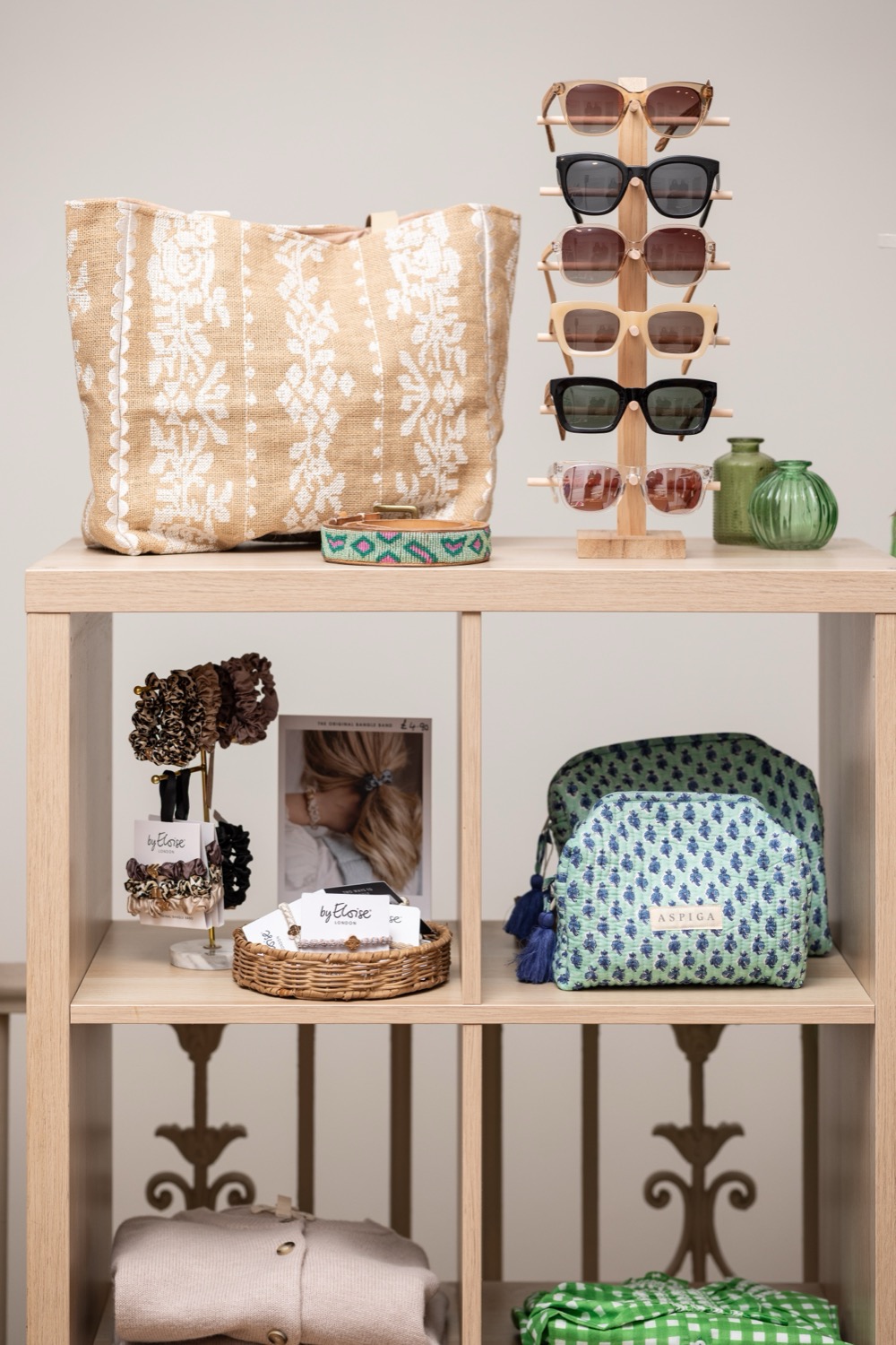 Sunglasses on a wooden display stand with woven tote bag, block-print wash bag and accessories on an oak shelf unit at Aspiga Kings Road London, retail product photography