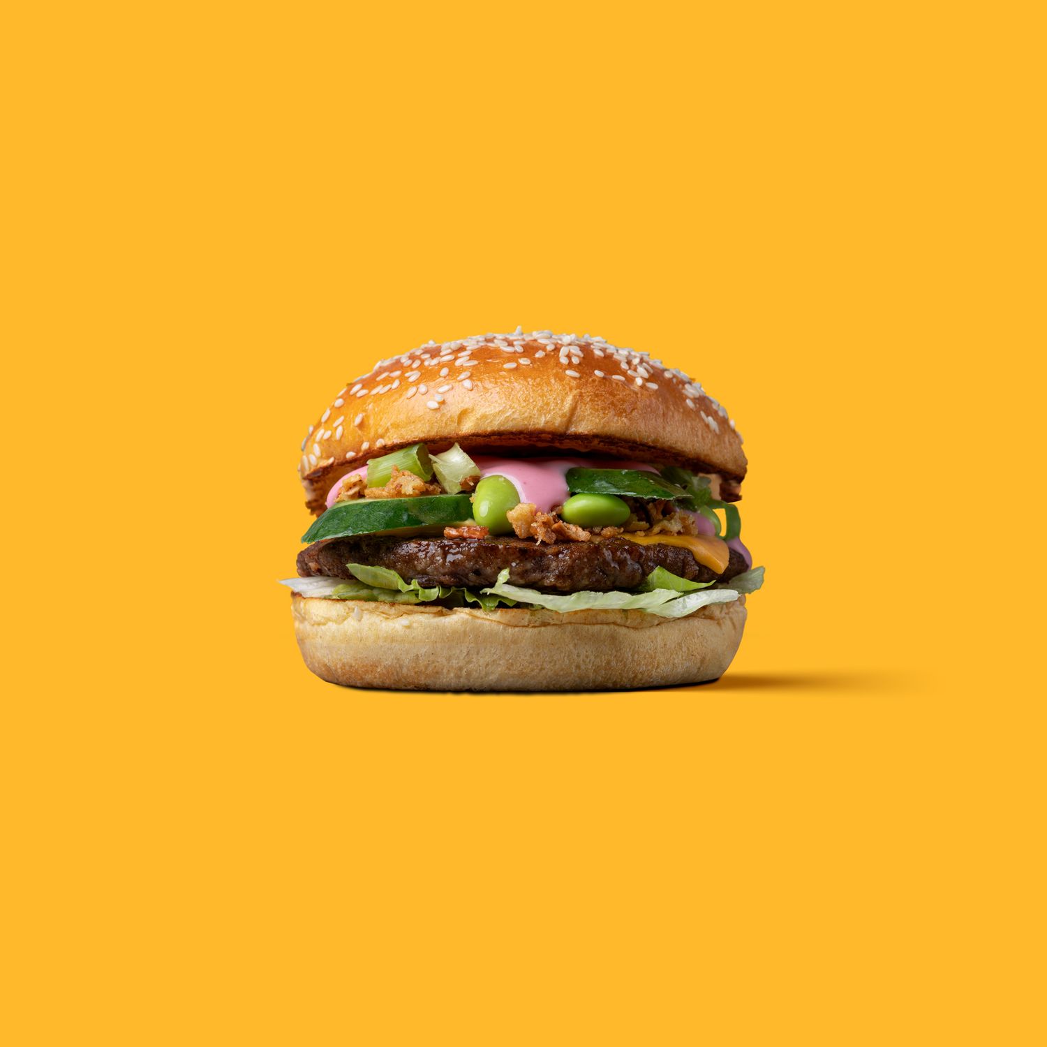 Gourmet burger with sesame bun, edamame, pink sauce and fresh toppings on a bright yellow background, clean studio food product photography Southampton