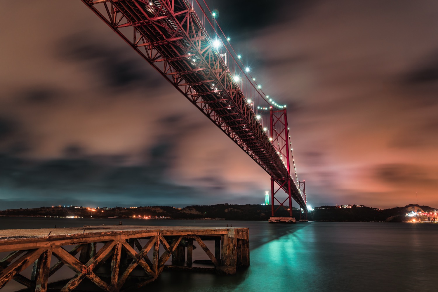 The 25 de Abril bridge at night from below, a long exposure with the lit deck reflected in the water, Lisbon, Portugal, travel photography