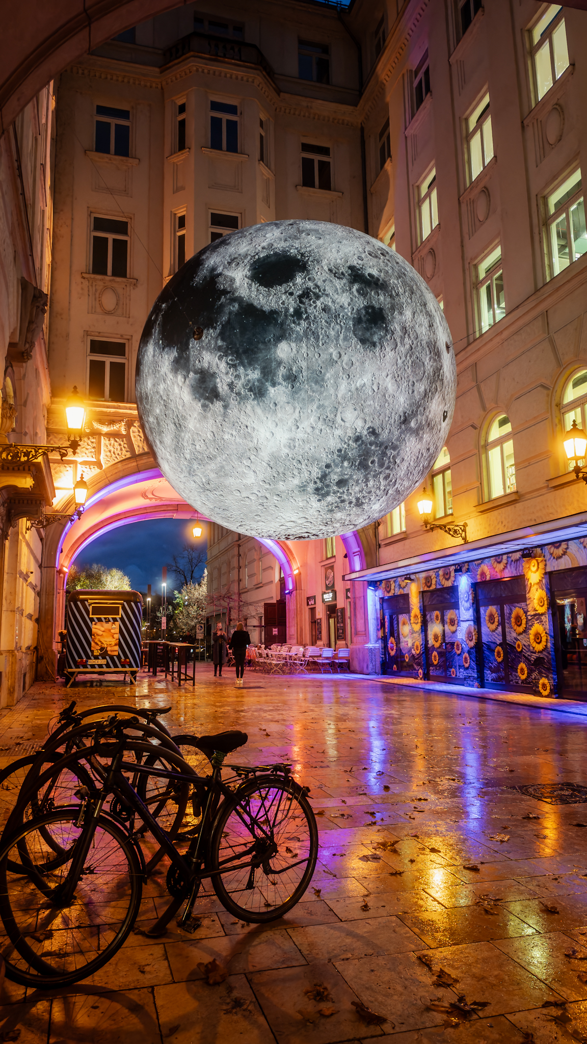 Neon-lit street at night with an illuminated moon installation overhead and bicycles on wet cobbles, Budapest, Hungary, travel photography