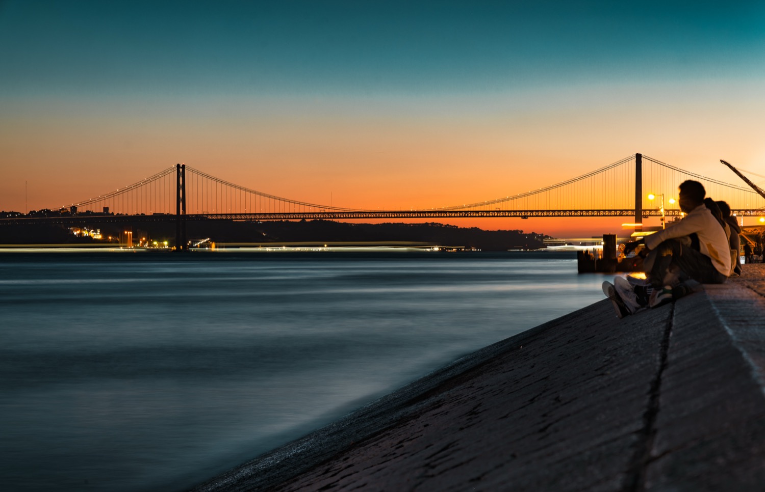 Sunset over the Tagus with the 25 de Abril bridge and a figure sitting on the river wall, Lisbon, Portugal, travel photography