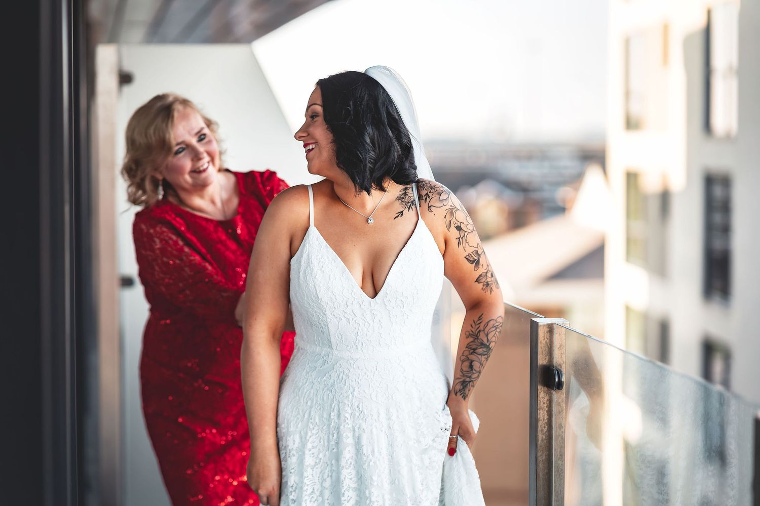 Tattooed bride and bridesmaid in red dress laughing together on a balcony at the Harbour Hotel Southampton during preparations, candid bridal prep photography