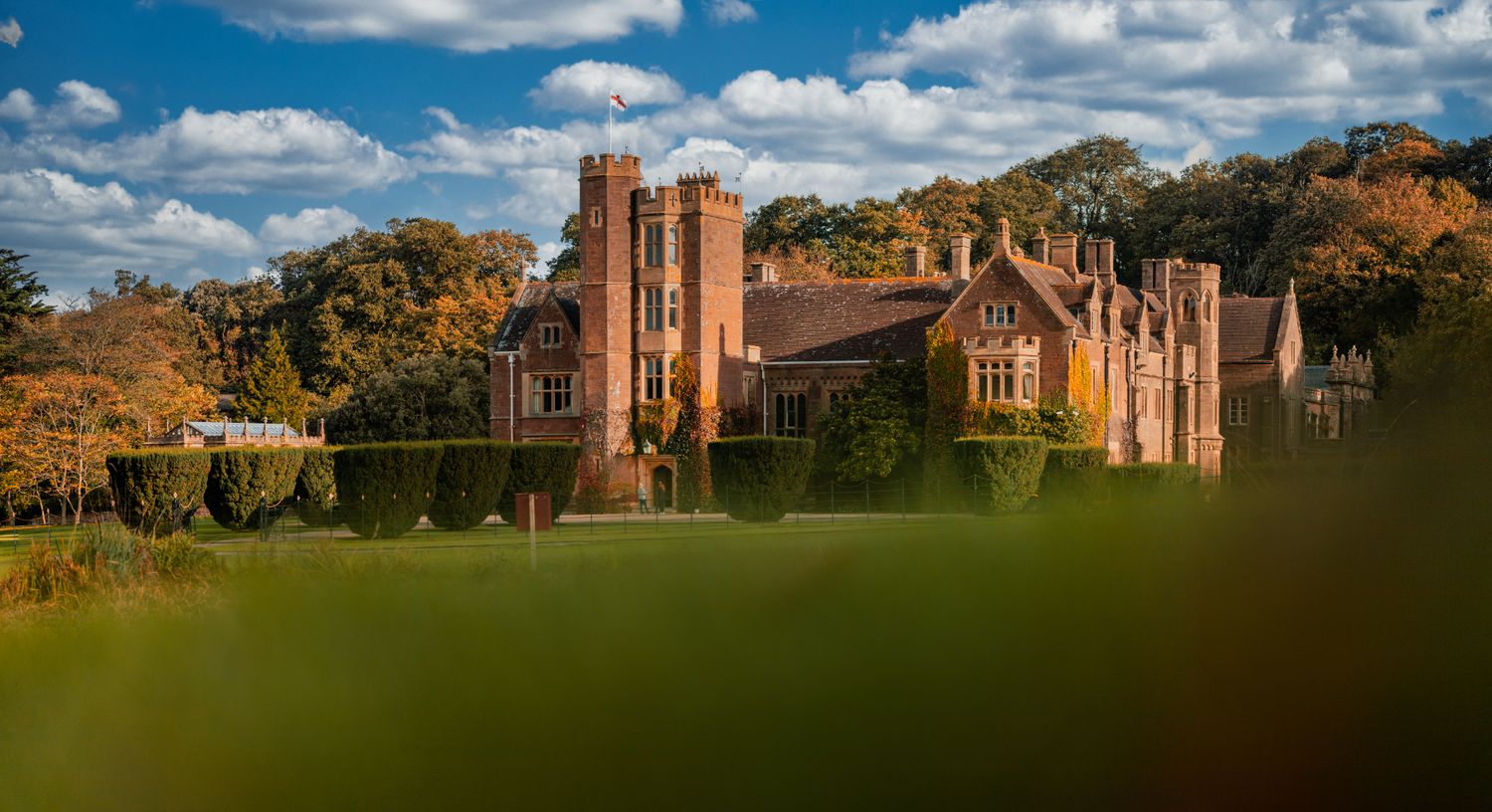 Red-brick Tudor manor wedding venue with a clock tower, framed by autumn trees and manicured lawns under a clear blue sky