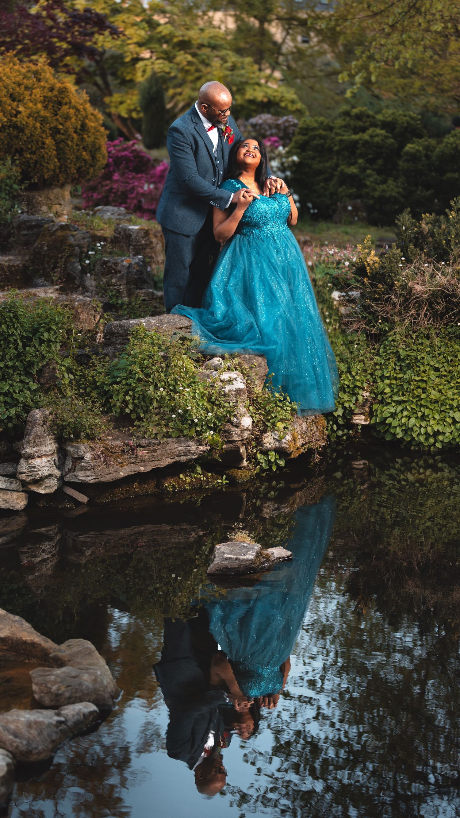 Couple standing on rocks by a garden pond with water reflection, bride in turquoise dress, romantic wedding photography Southampton