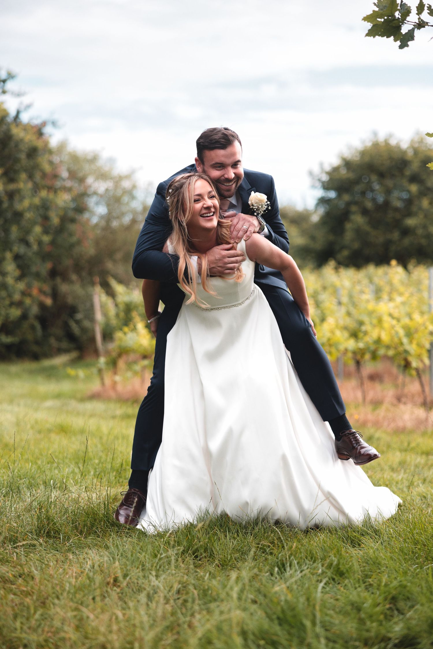 Groom wrapping his arms around the bride from behind on a sunlit lawn with vineyard countryside beyond, relaxed natural wedding photography