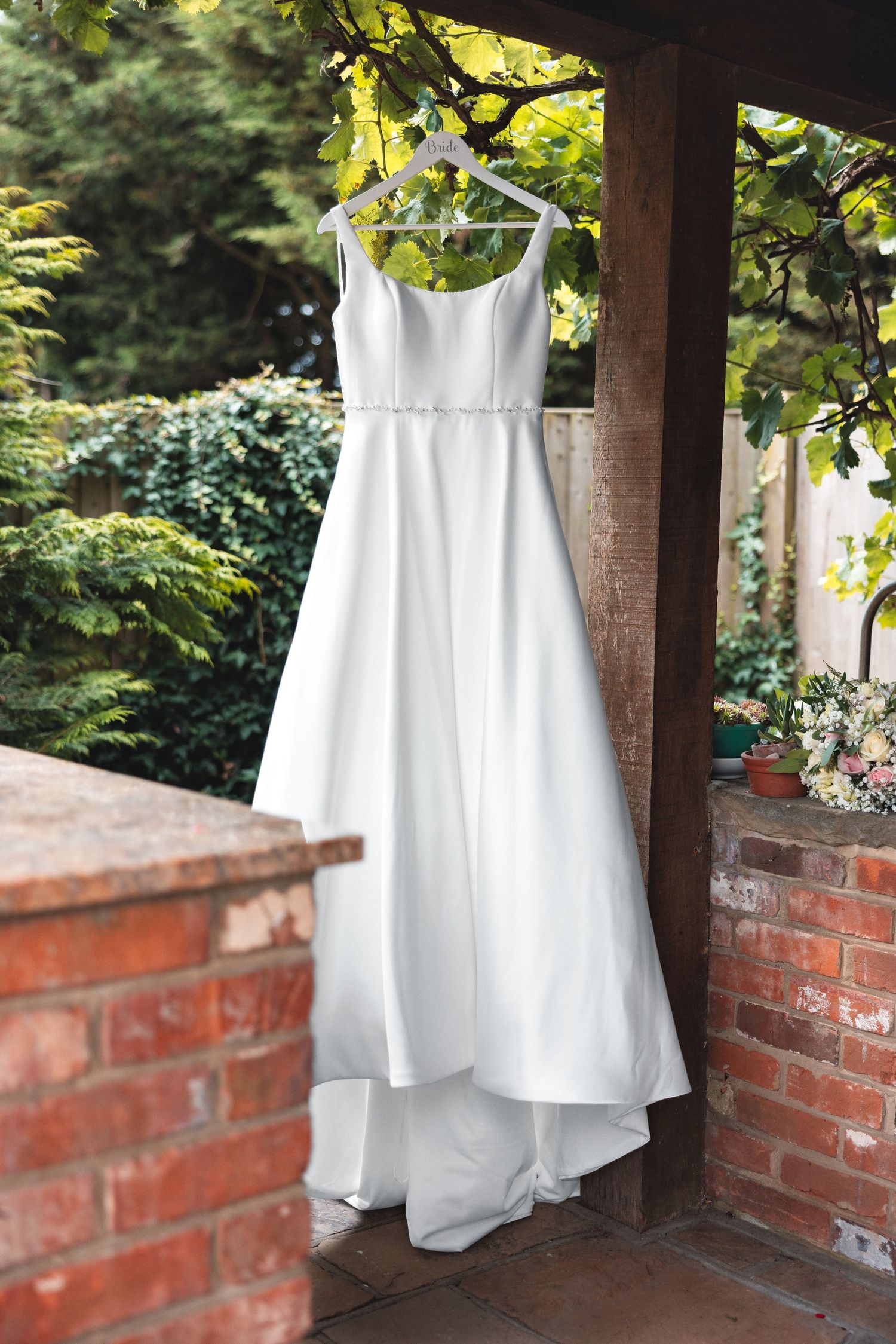 Wedding dress hanging on a wooden garden pergola beam with green foliage behind, bridal detail photography Southampton