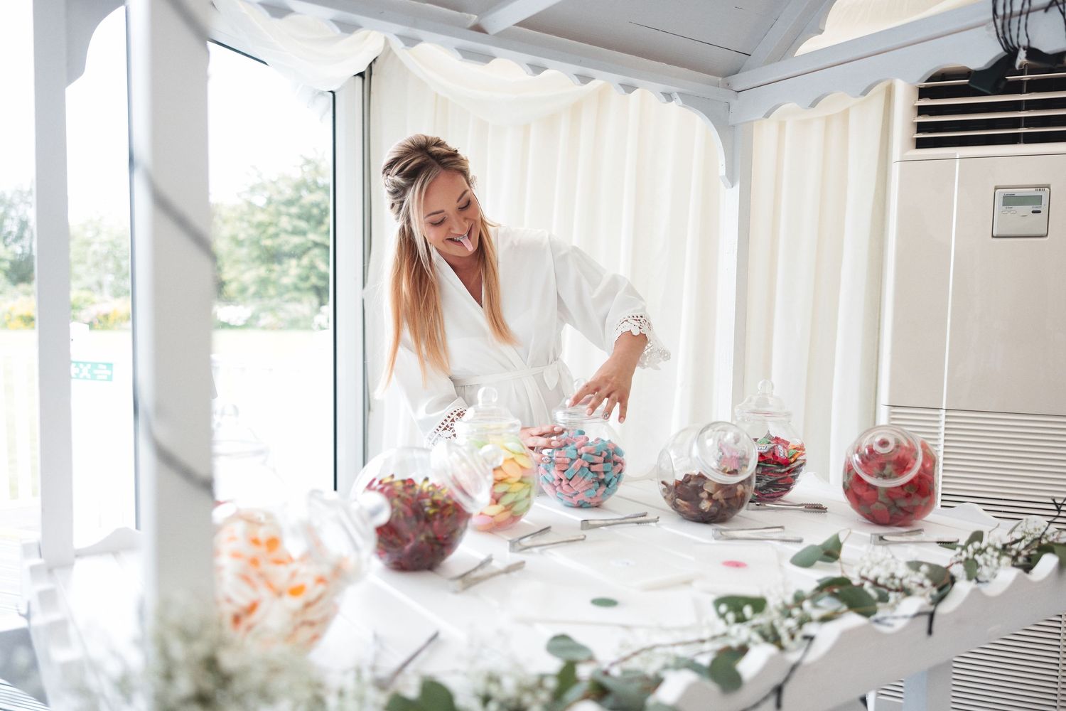 Bride in white robe picking sweets from a candy table with colourful glass jars at the wedding venue, candid wedding moment photography Southampton