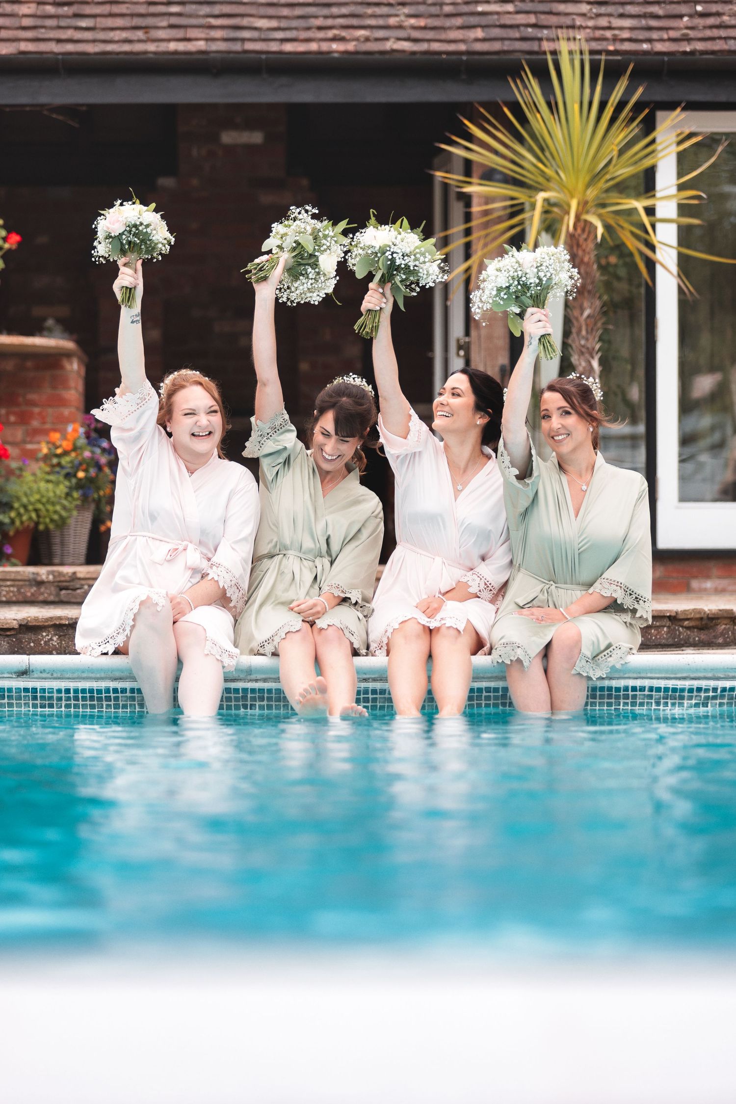 Bridesmaids in matching robes sitting along the edge of a swimming pool with their feet in the water, raising their bouquets, fun candid wedding photography