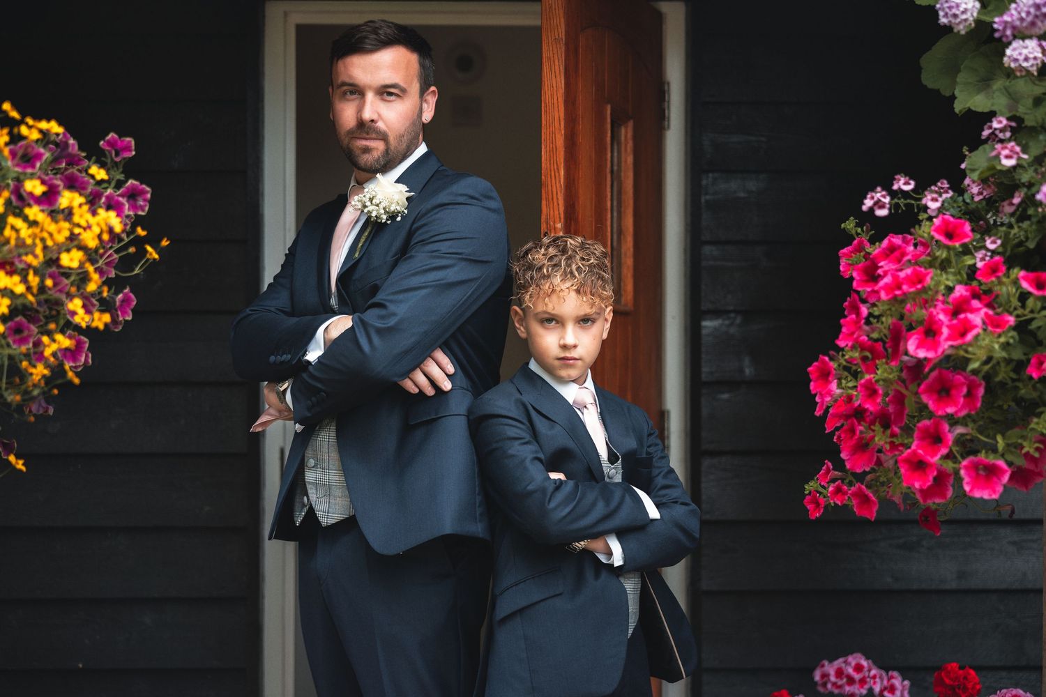 Groom in navy suit standing with young page boy in matching suit with arms crossed by a doorway with hanging flower baskets, wedding portrait photography Southampton
