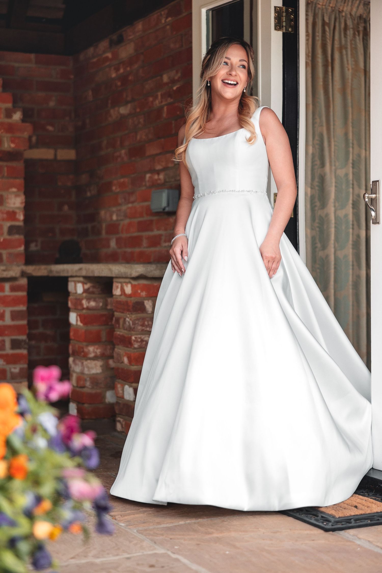 Bride smiling in a white A-line dress standing in a brick doorway with colourful flowers in foreground, bridal portrait photography Southampton