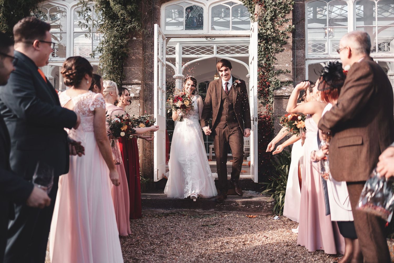 Bride and groom walking through confetti outside a stone manor house with bridesmaids in pink, joyful wedding exit photography St Audrie\