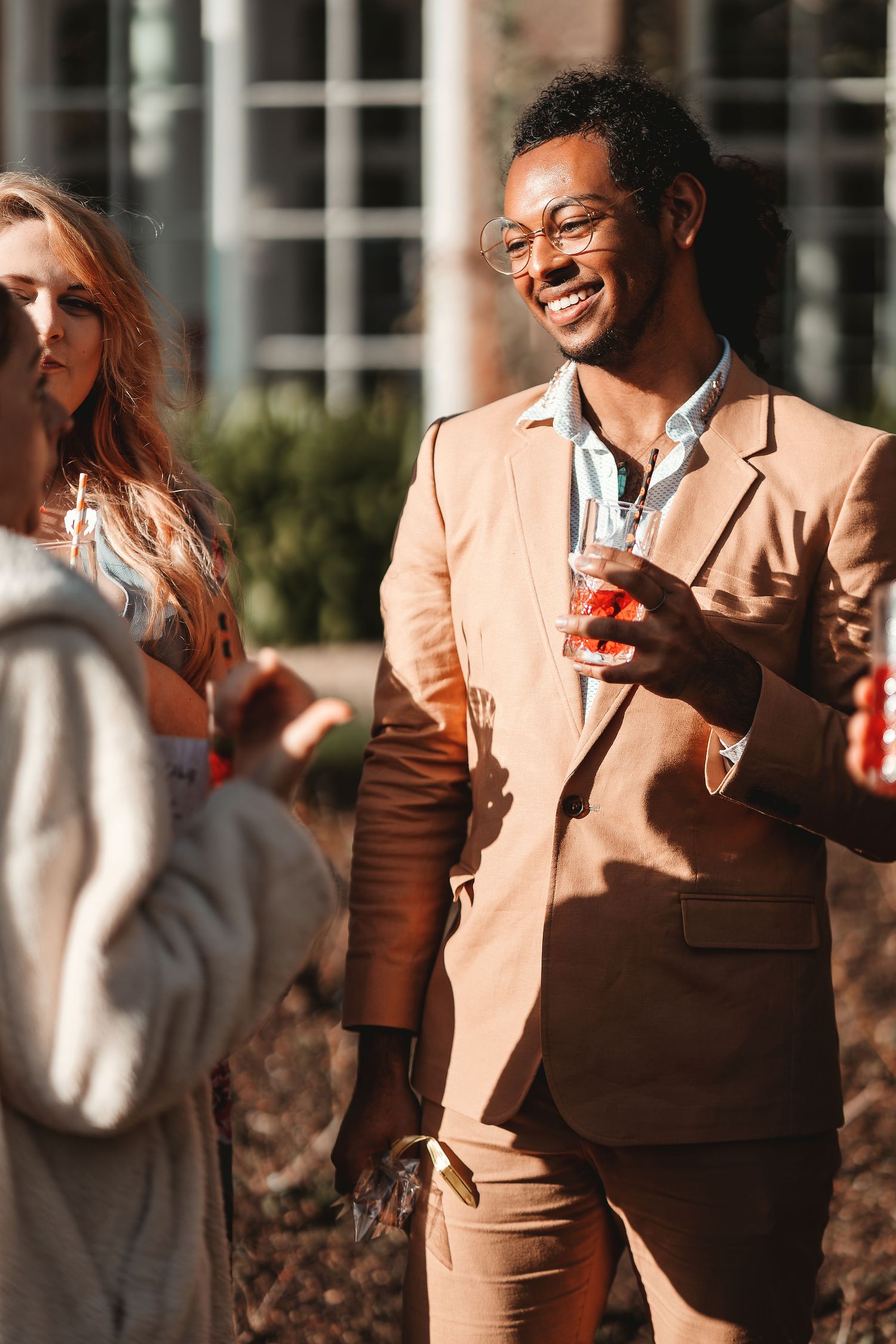 Smartly dressed wedding guest in a tan suit with glasses holding a drink and chatting outdoors in sunshine, candid wedding guest photography St Audrie\