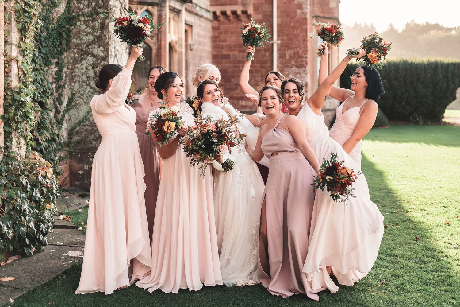 Bride and bridesmaids in blush pink dresses raising their autumn bouquets outside a stone manor house, candid group wedding photography
